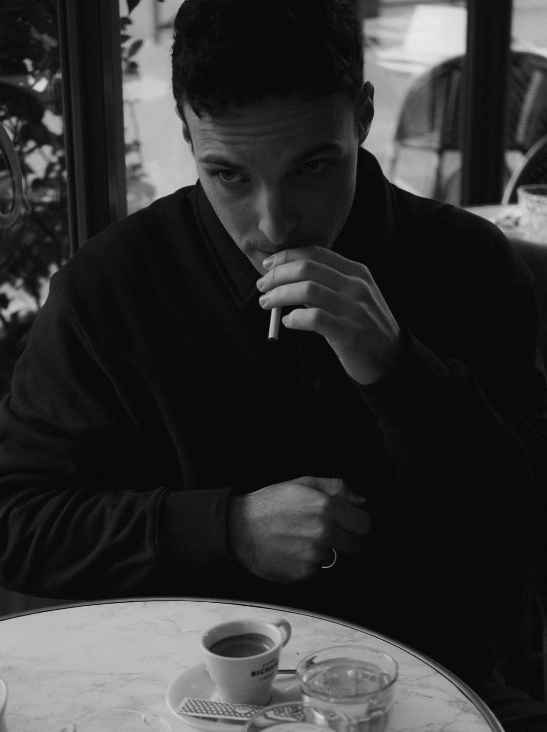 Black and white photo of a young man lighting a cigarette at a cafe table with a coffee cup.