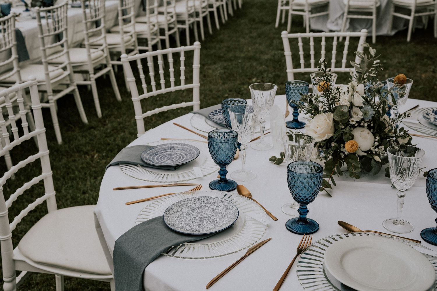 Elegant outdoor wedding reception table setting with blue goblets, gray plates, gold cutlery, and floral centerpiece on white linen.