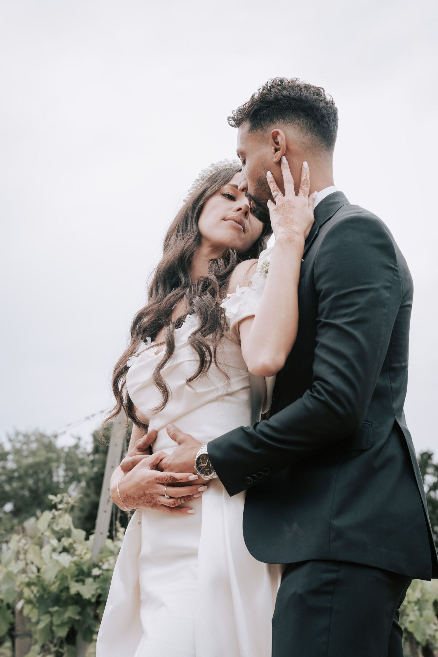 Bride tenderly touches grooms face as they embrace in vineyard setting under cloudy sky.