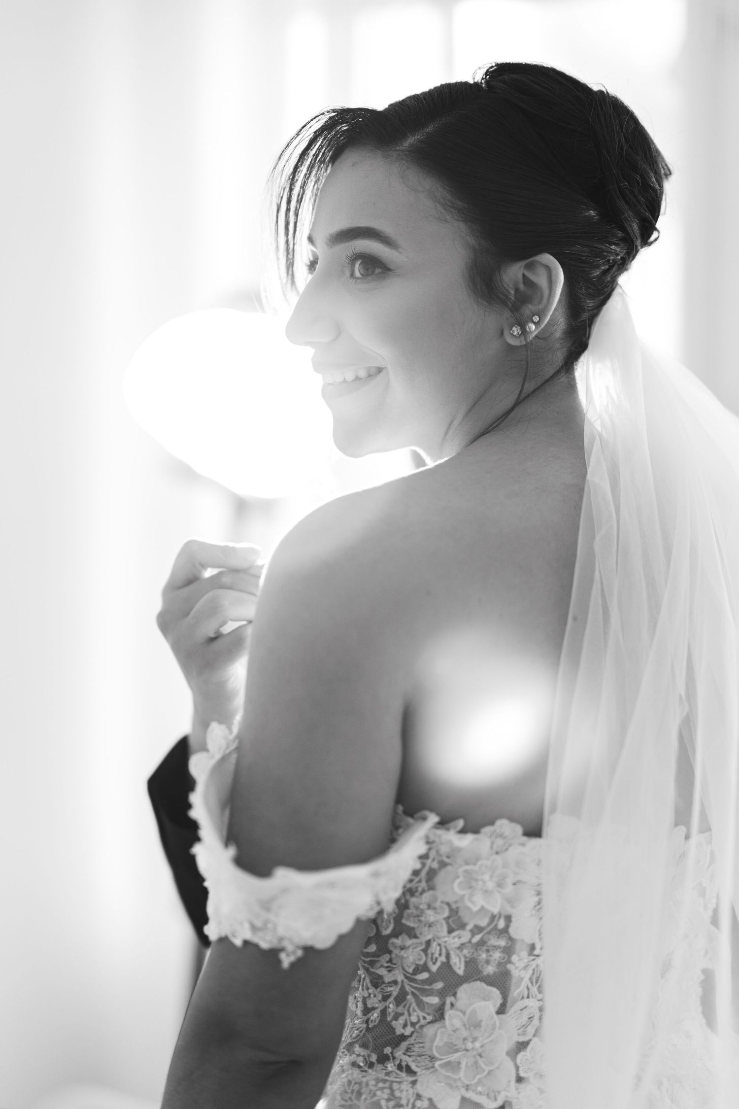 Black and white portrait of smiling bride in lace gown and veil by window.
