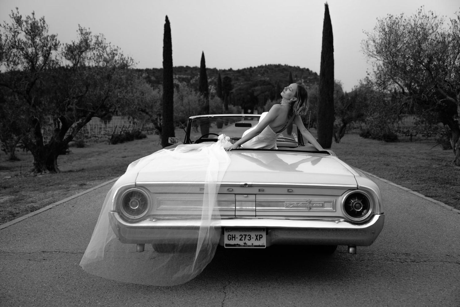 Bride in strapless gown with flowing veil poses dramatically on a white vintage Ford convertible in black and white.