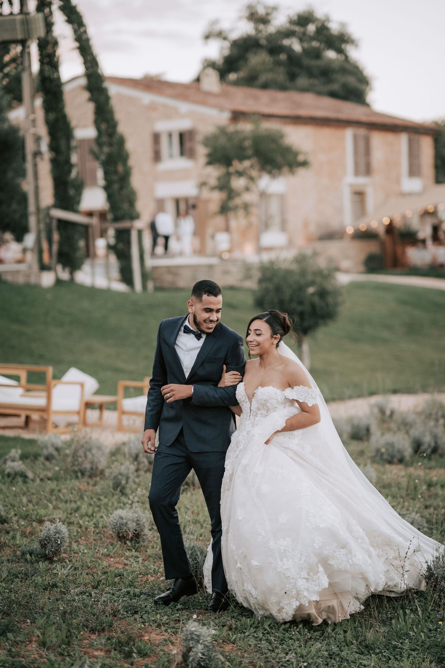 Smiling newlyweds walk arm in arm across garden lawn at rustic stone chateau wedding venue.