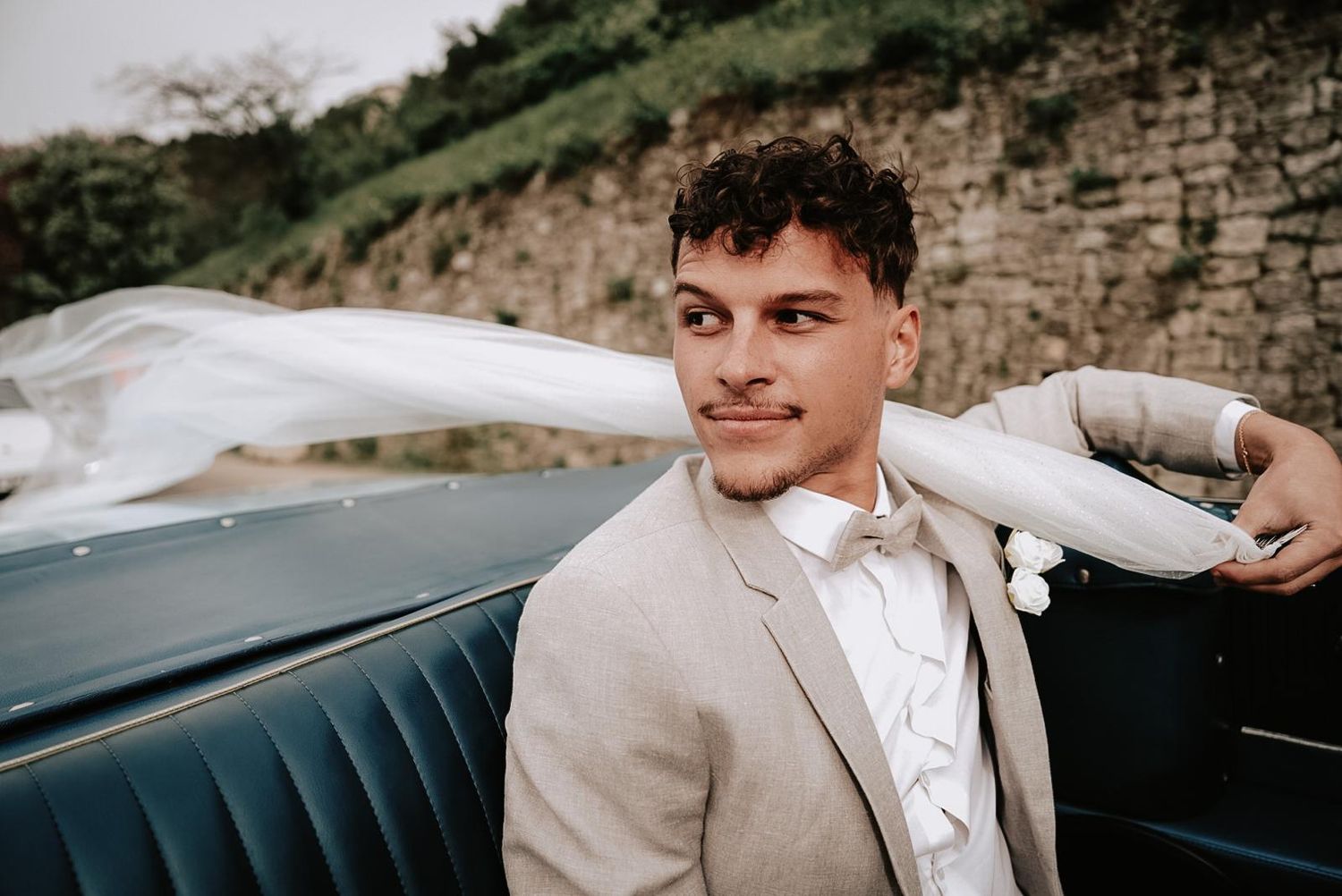 Handsome groom in beige suit and bow tie smiles while leaning on a vintage blue convertible car.