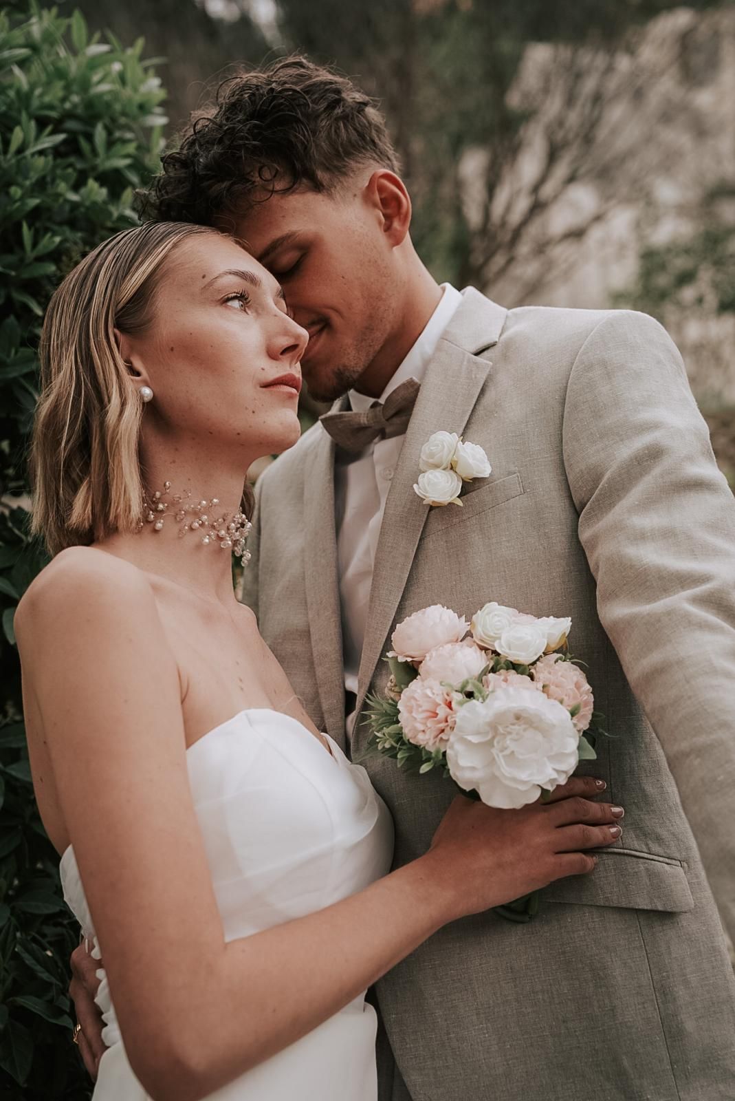 Bride and groom share an intimate moment, she in a strapless white gown holding pink peonies, he in a beige linen suit.