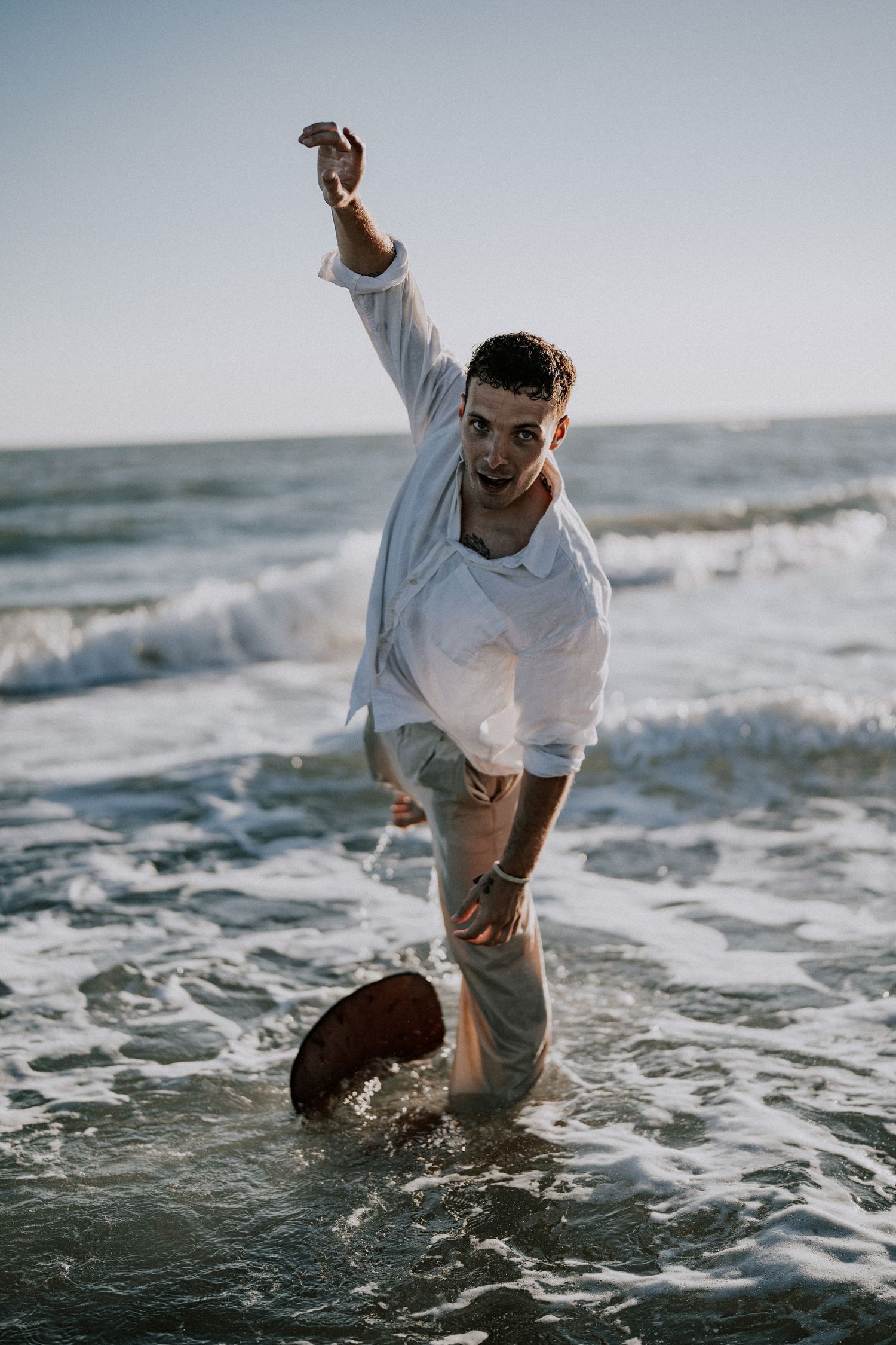 A wet young man in a white shirt stands in ocean waves with one arm raised at the beach.