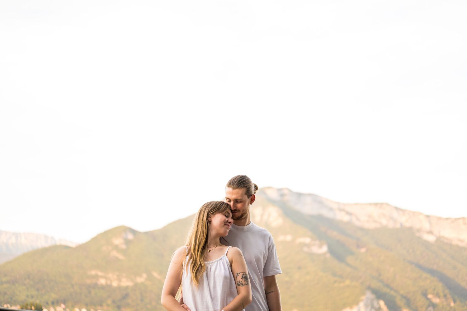 Couple standing together with mountain backdrop at golden hour, woman leaning her head against mans shoulder.