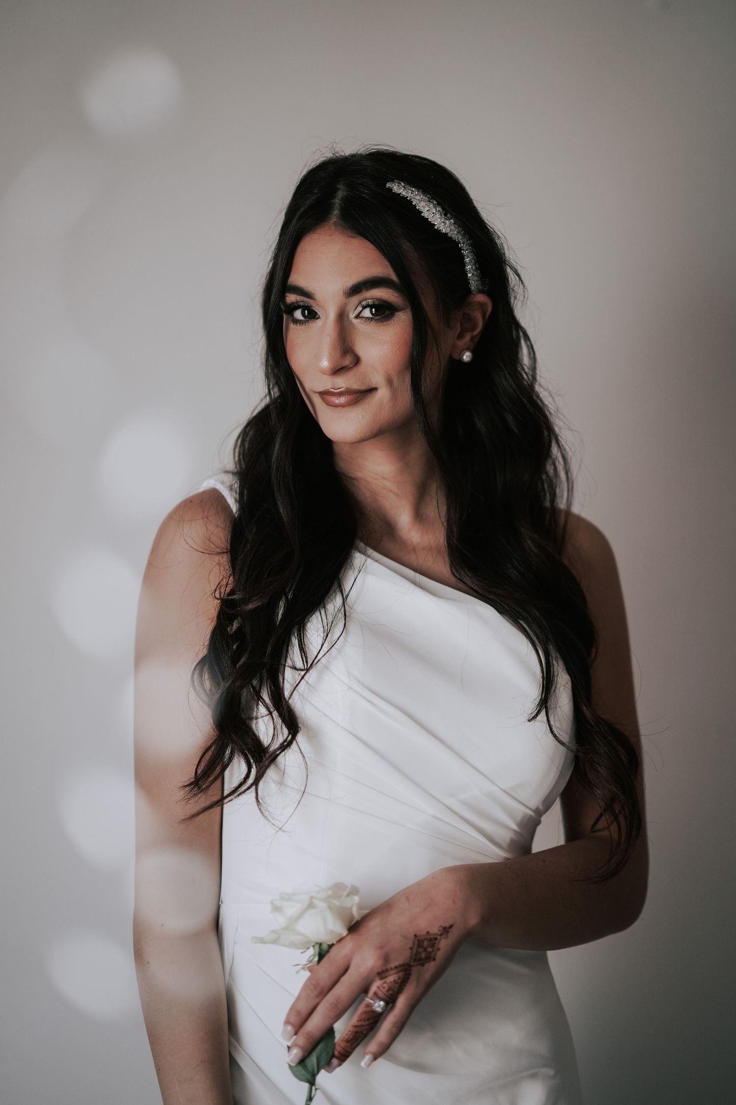 Elegant bride in white gown holding a flower, wearing a crystal headband and tattoos.