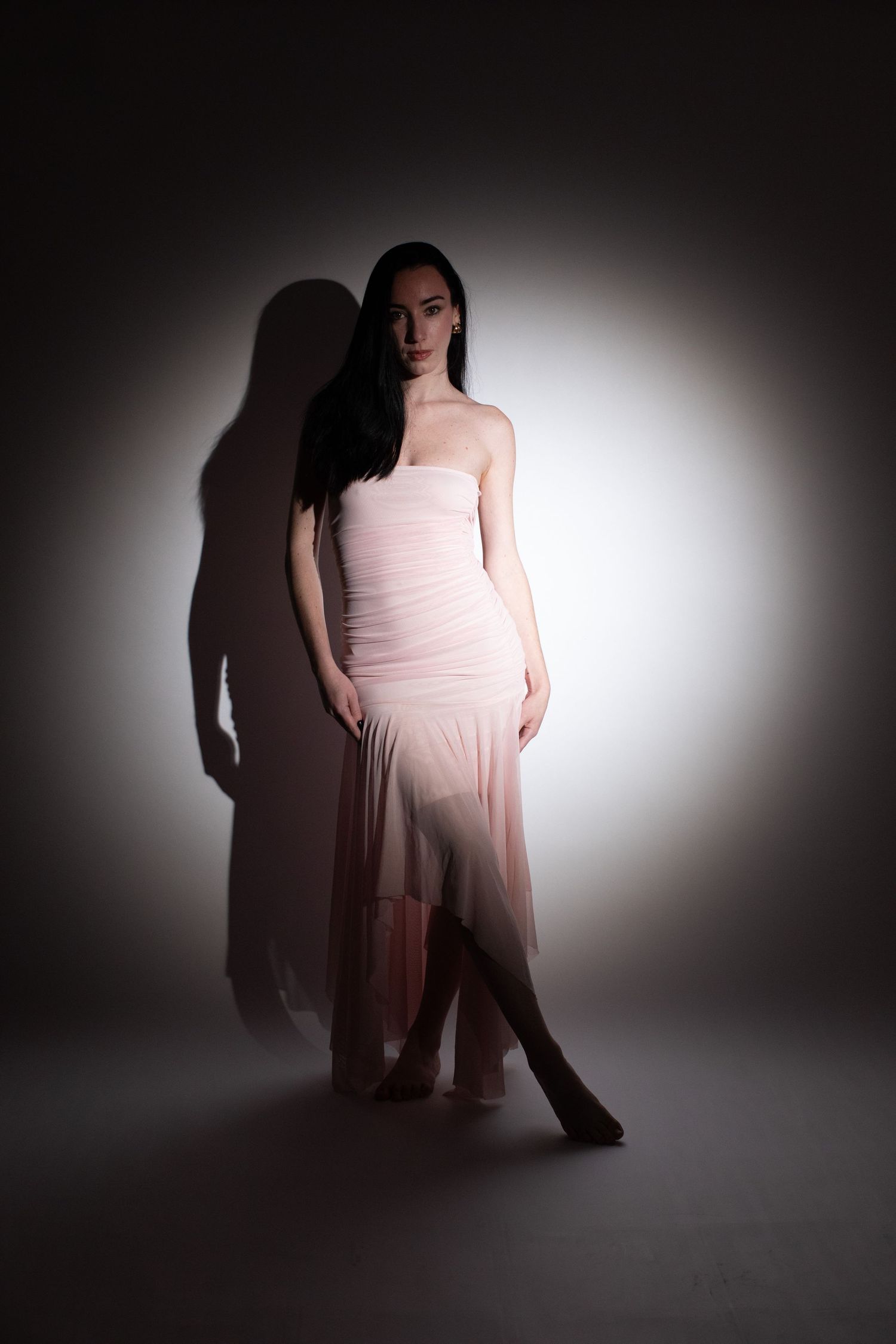 Young woman in a strapless pink chiffon dress posing dramatically against a dark studio background with spotlight.