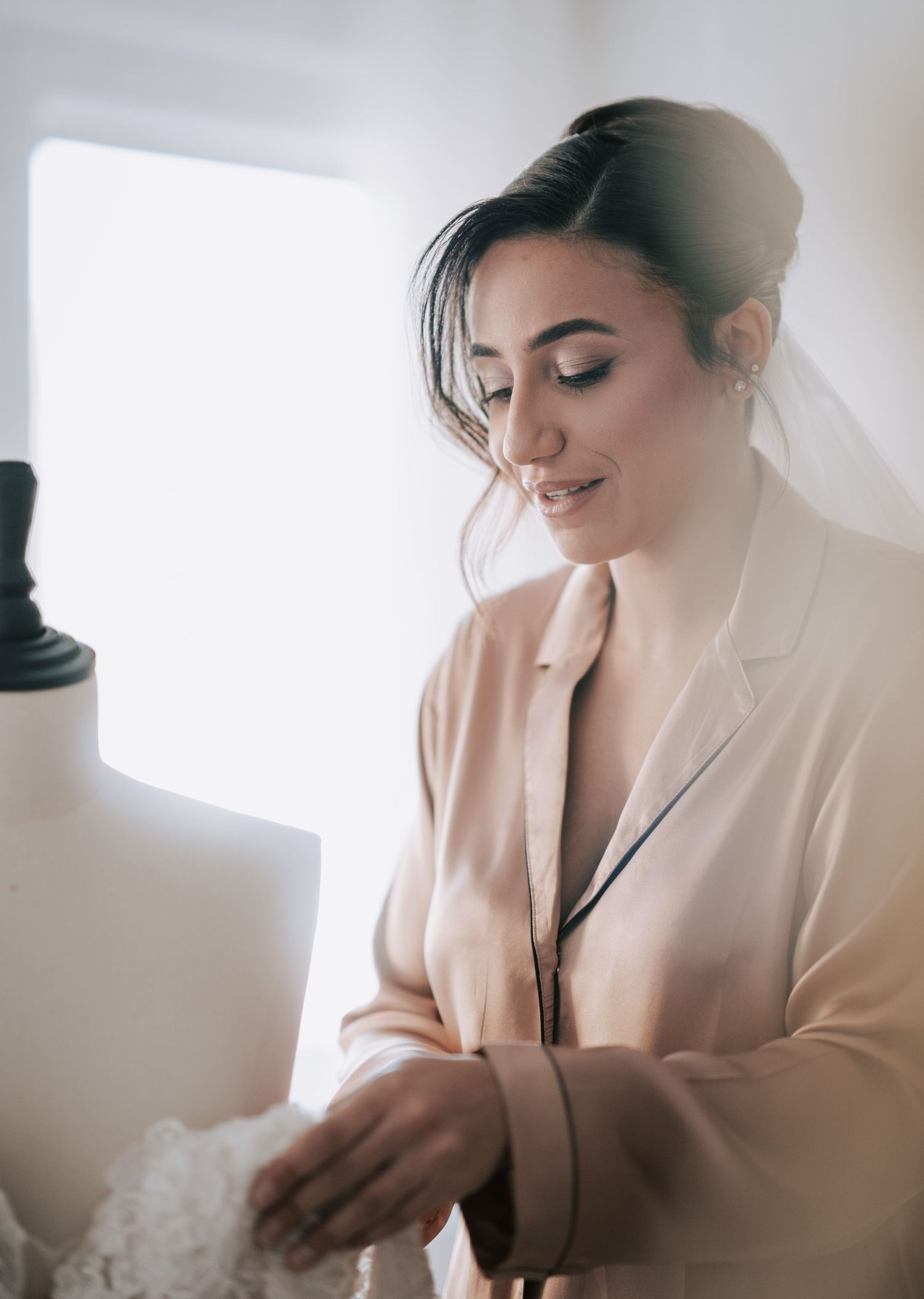 Elegant bride in champagne silk robe smiling while preparing wedding dress on mannequin.