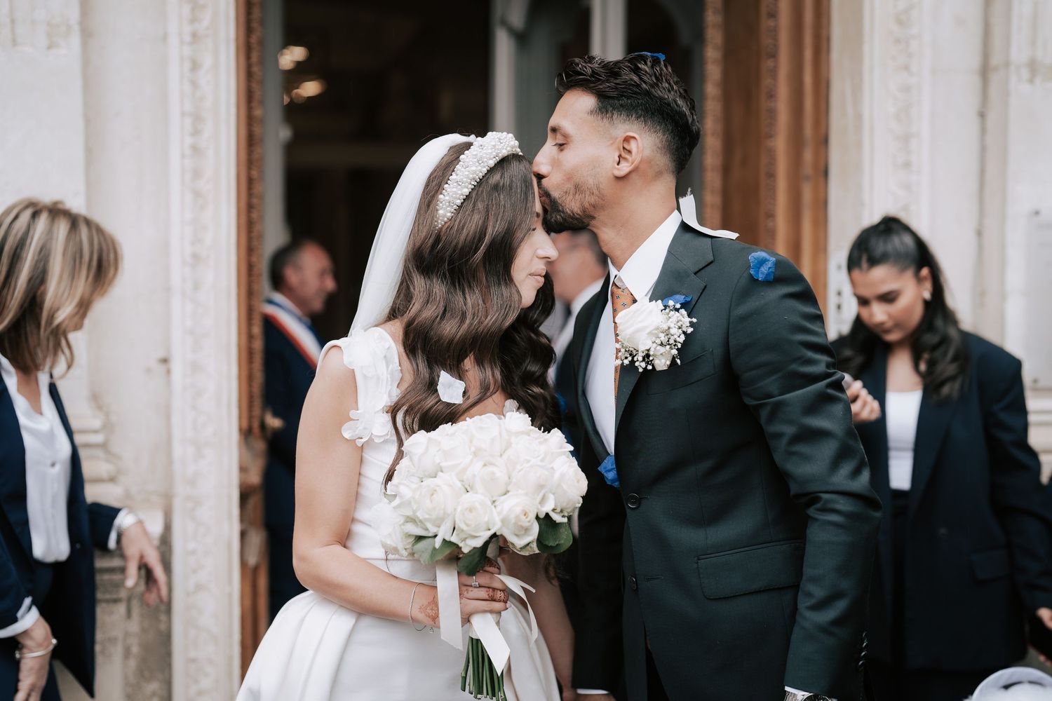 Groom kisses bride outside ornate church doors as guests gather in background.