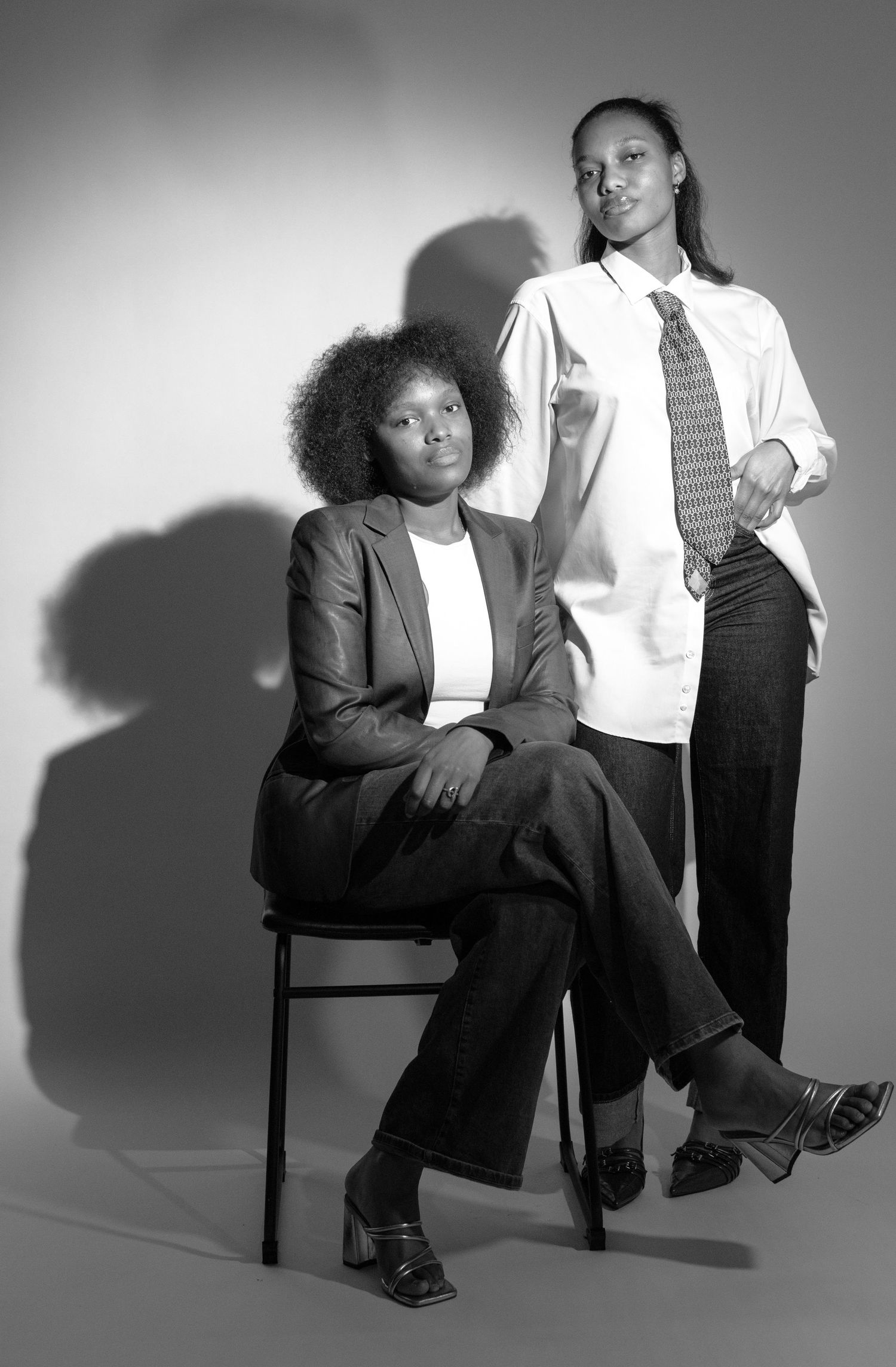 Black and white portrait of two young women posing confidently, one seated in a chair, both wearing business casual attire.