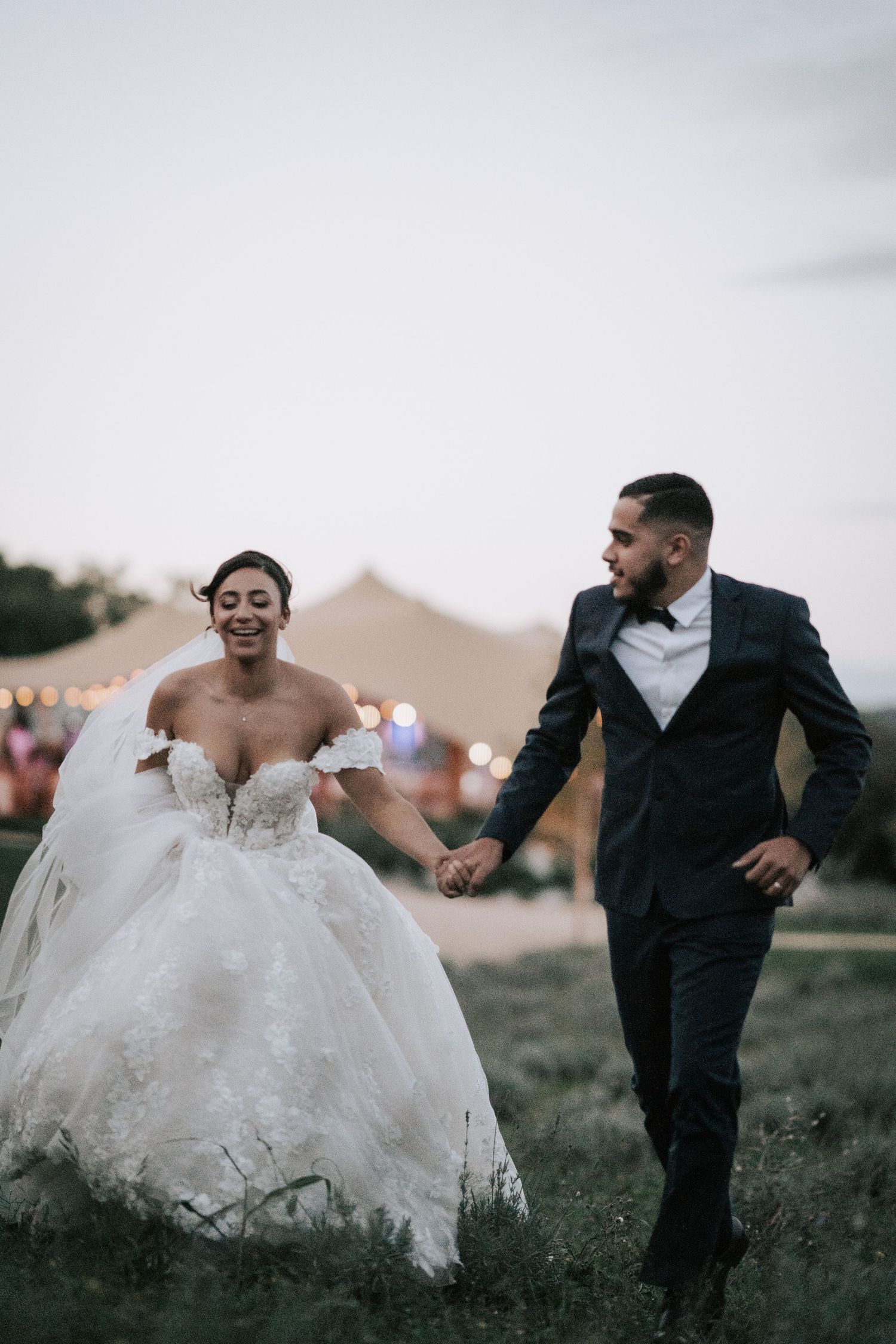 Joyful bride and groom holding hands and running through a field at dusk with a wedding tent behind them.