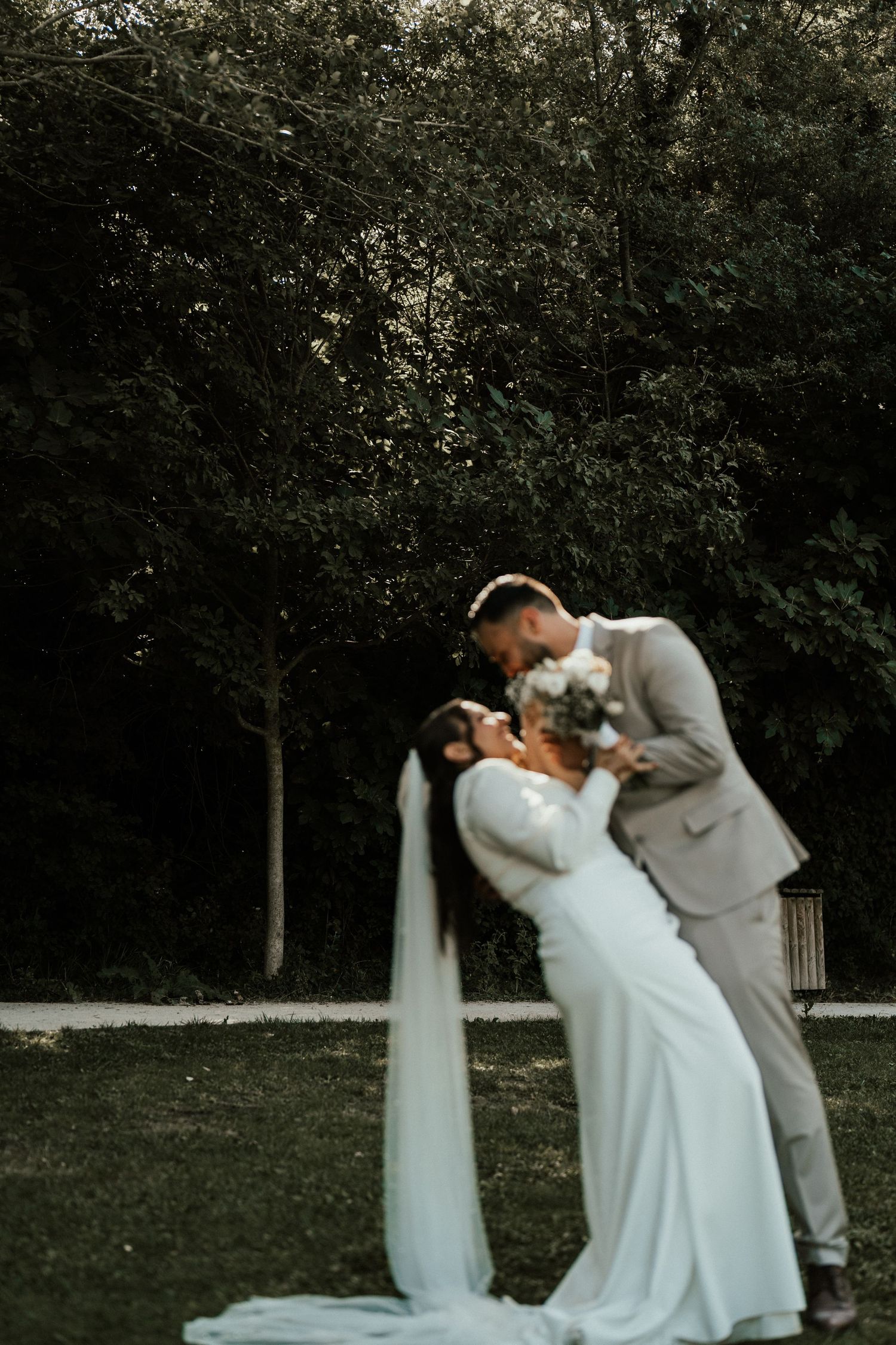 Groom in beige suit dips bride in white gown with long veil for romantic kiss in garden.