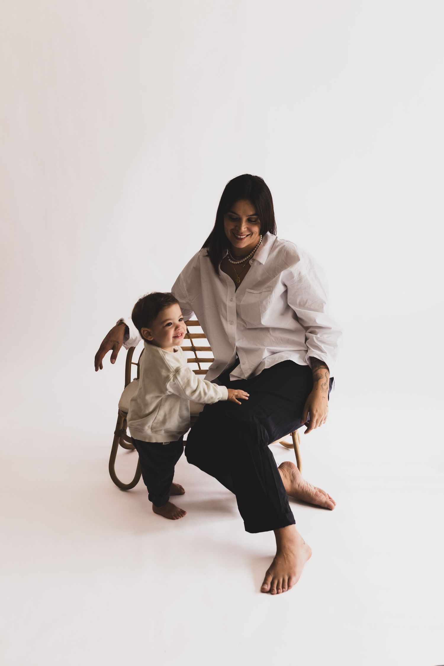Woman in white shirt sitting in a wooden chair smiling while a young toddler stands beside her in a studio.