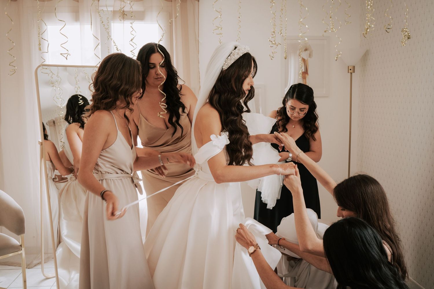 Bridesmaids in champagne gowns help bride into her white wedding dress in a bright dressing room.