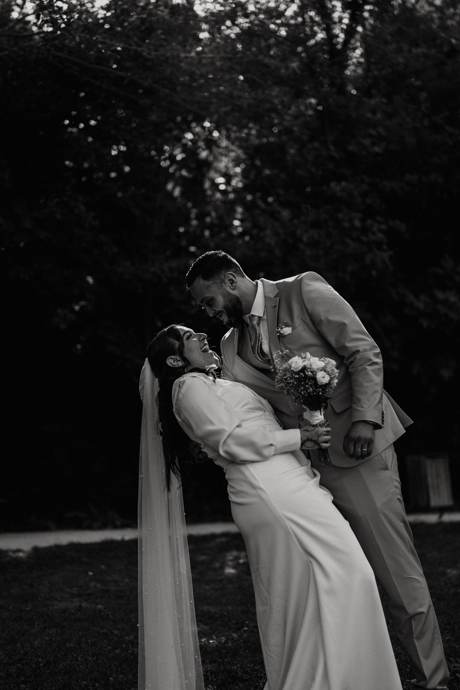 Black and white photo of laughing bride and groom sharing romantic dip kiss in lush garden setting.