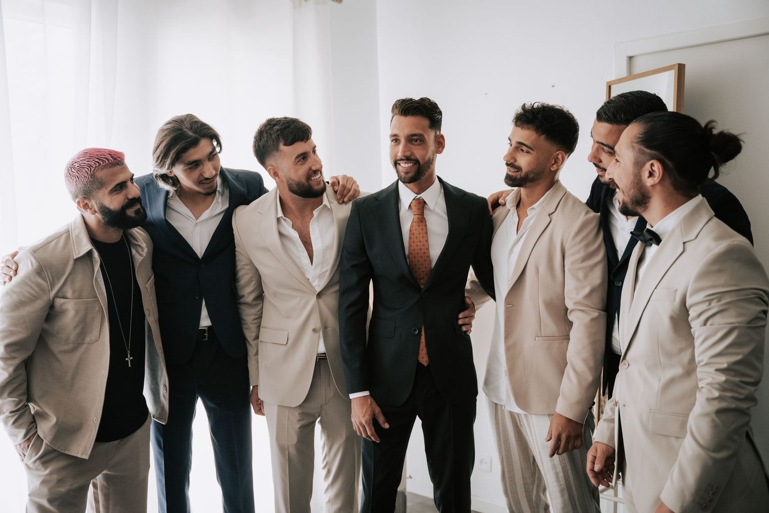 Groom in dark suit with groomsmen in beige suits smiling together before the wedding ceremony.