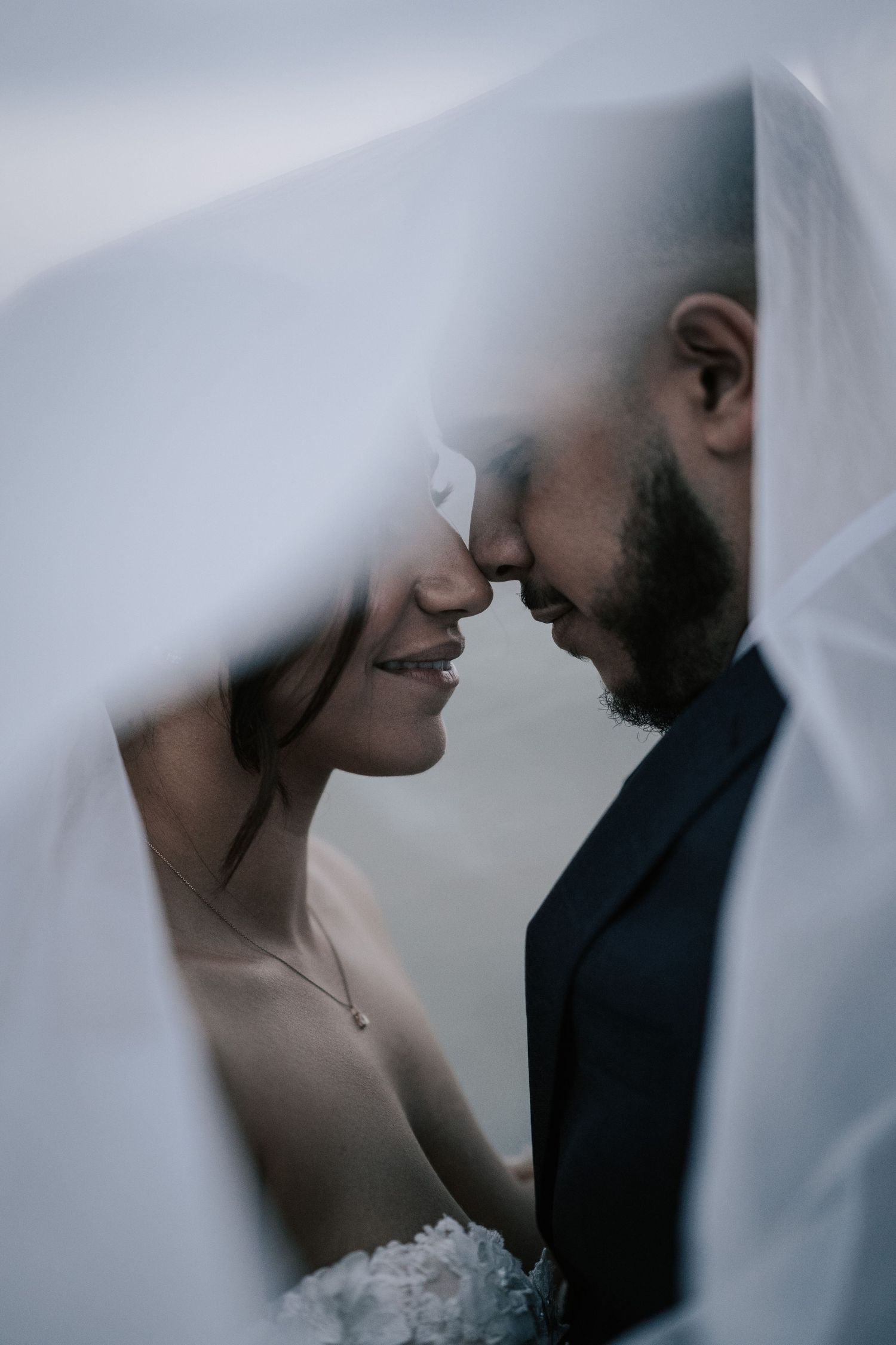 Bride and groom share an intimate moment under a flowing white veil, touching foreheads and smiling on their wedding day.