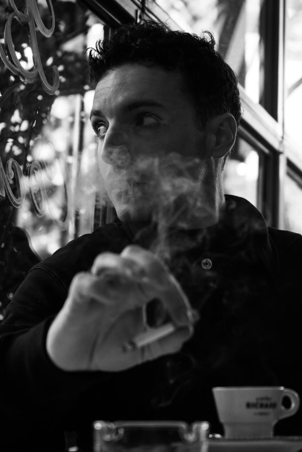 Black and white portrait of a man smoking a cigarette in a cafe, exhaling smoke dramatically.