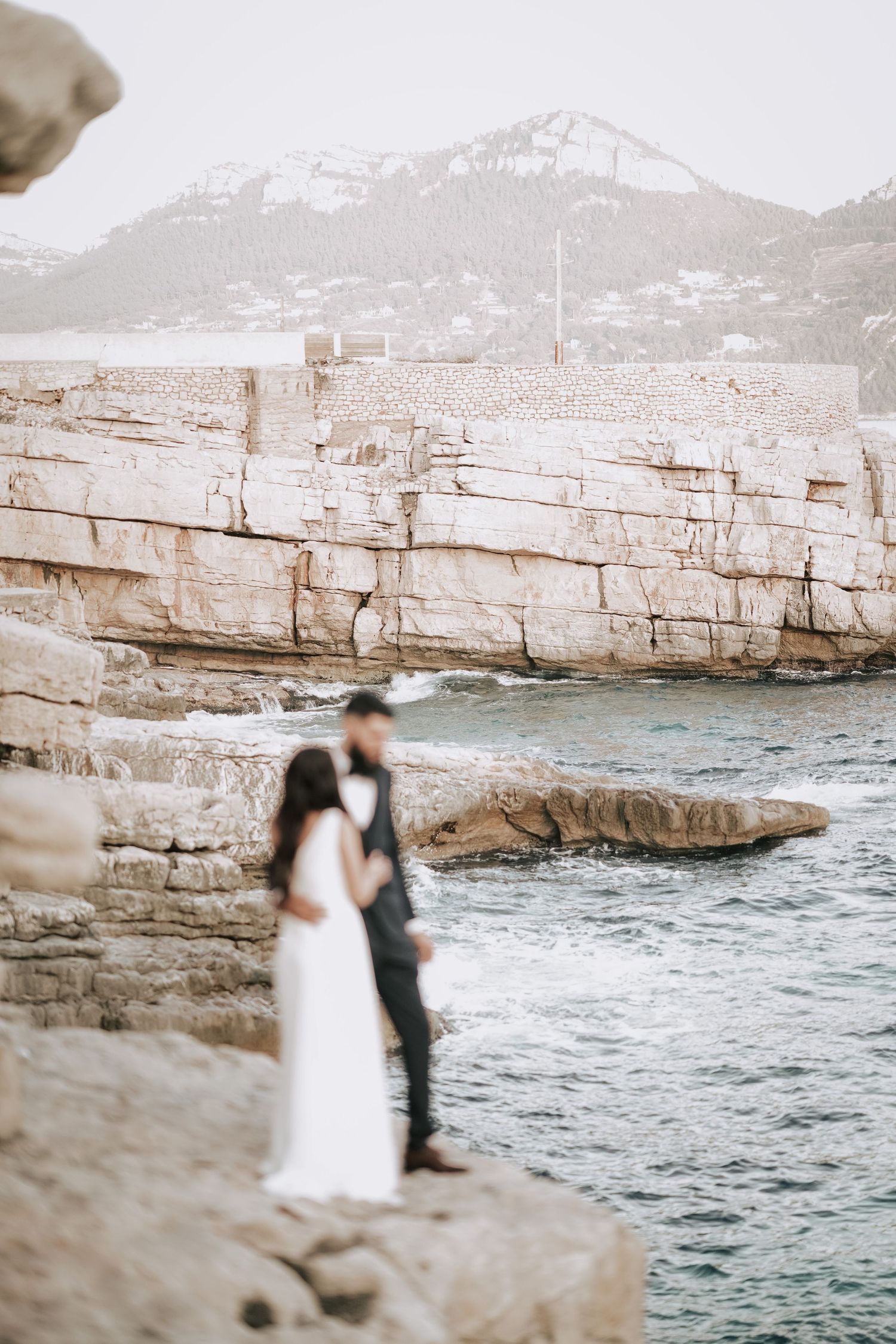 Bride and groom standing on dramatic rocky coastal cliffs with turquoise water and mountains behind.