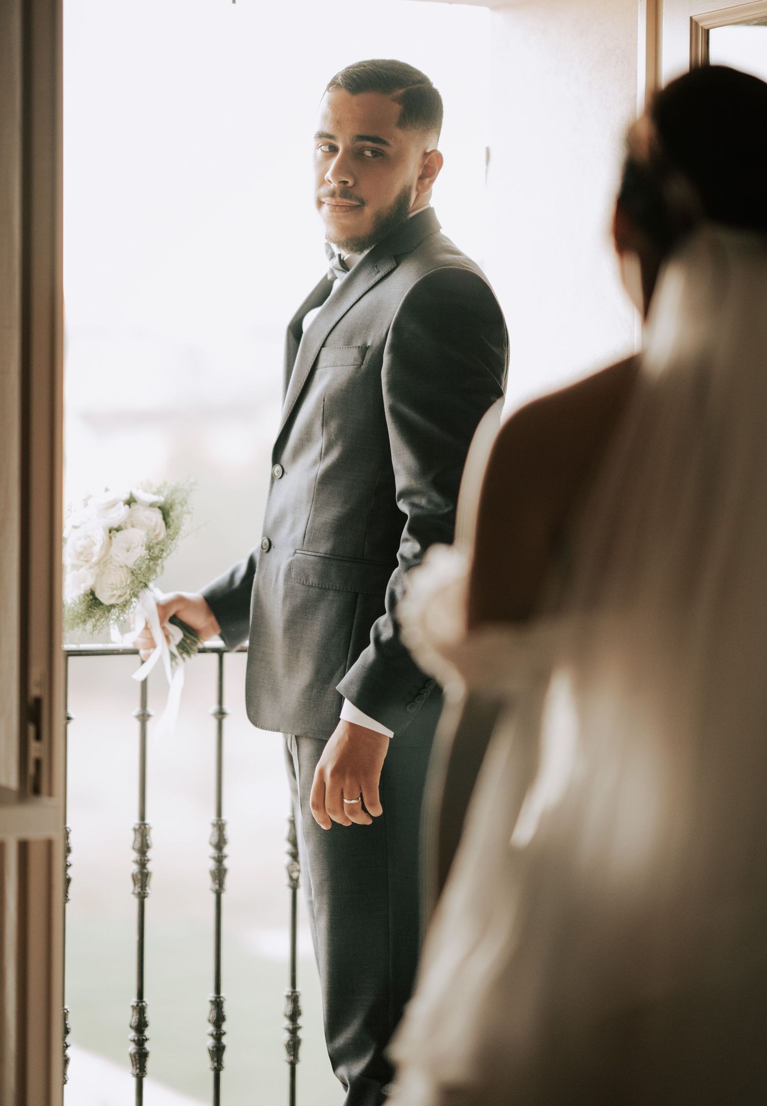 Groom in gray suit holds bouquet while seeing bride for the first time.