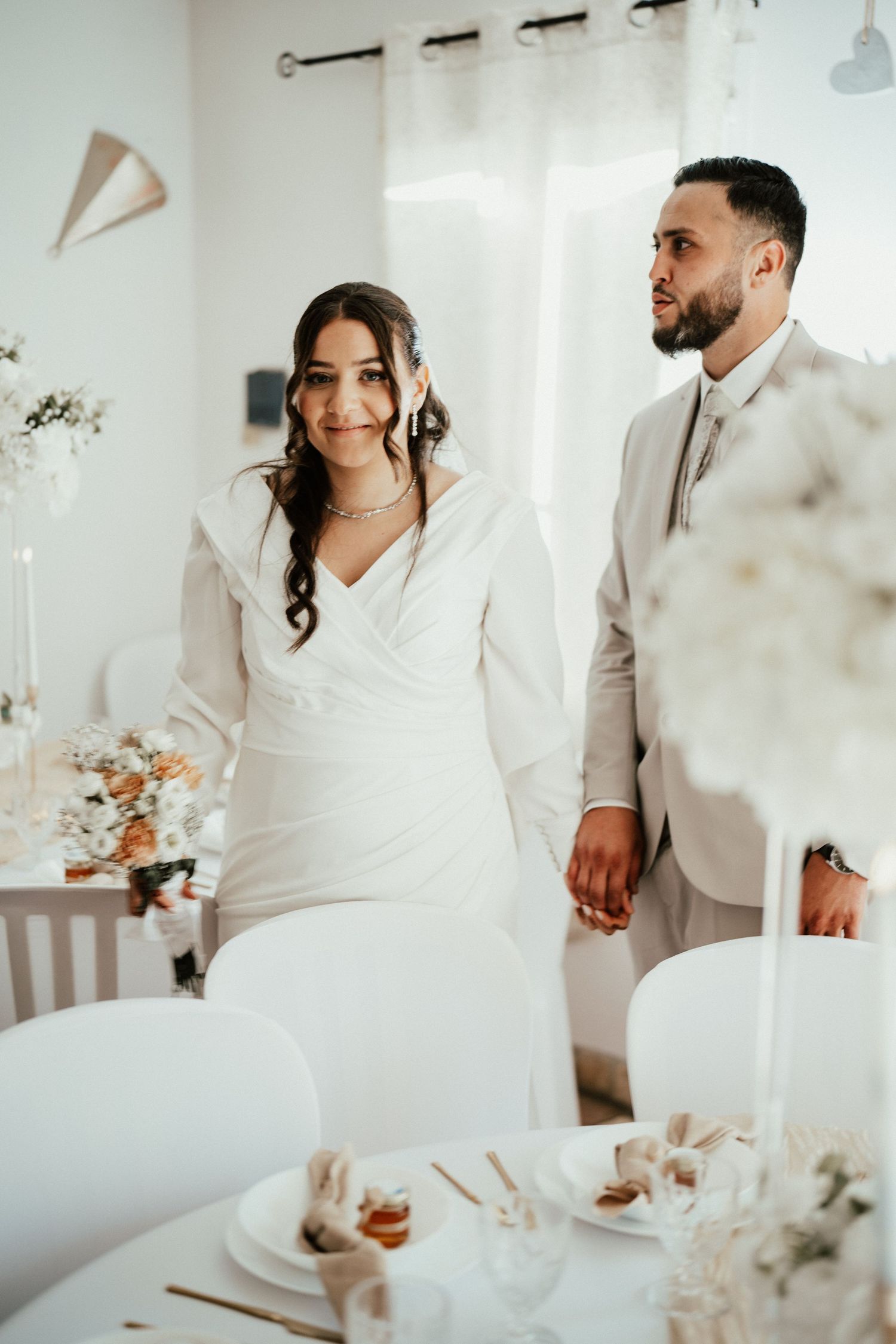 Bride in white long-sleeve gown and groom in beige suit hold hands at elegant white wedding reception.