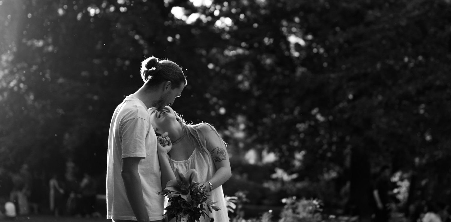 Black and white photo of a couple sharing an intimate moment outdoors, with the groom leaning toward the bride holding flowers.