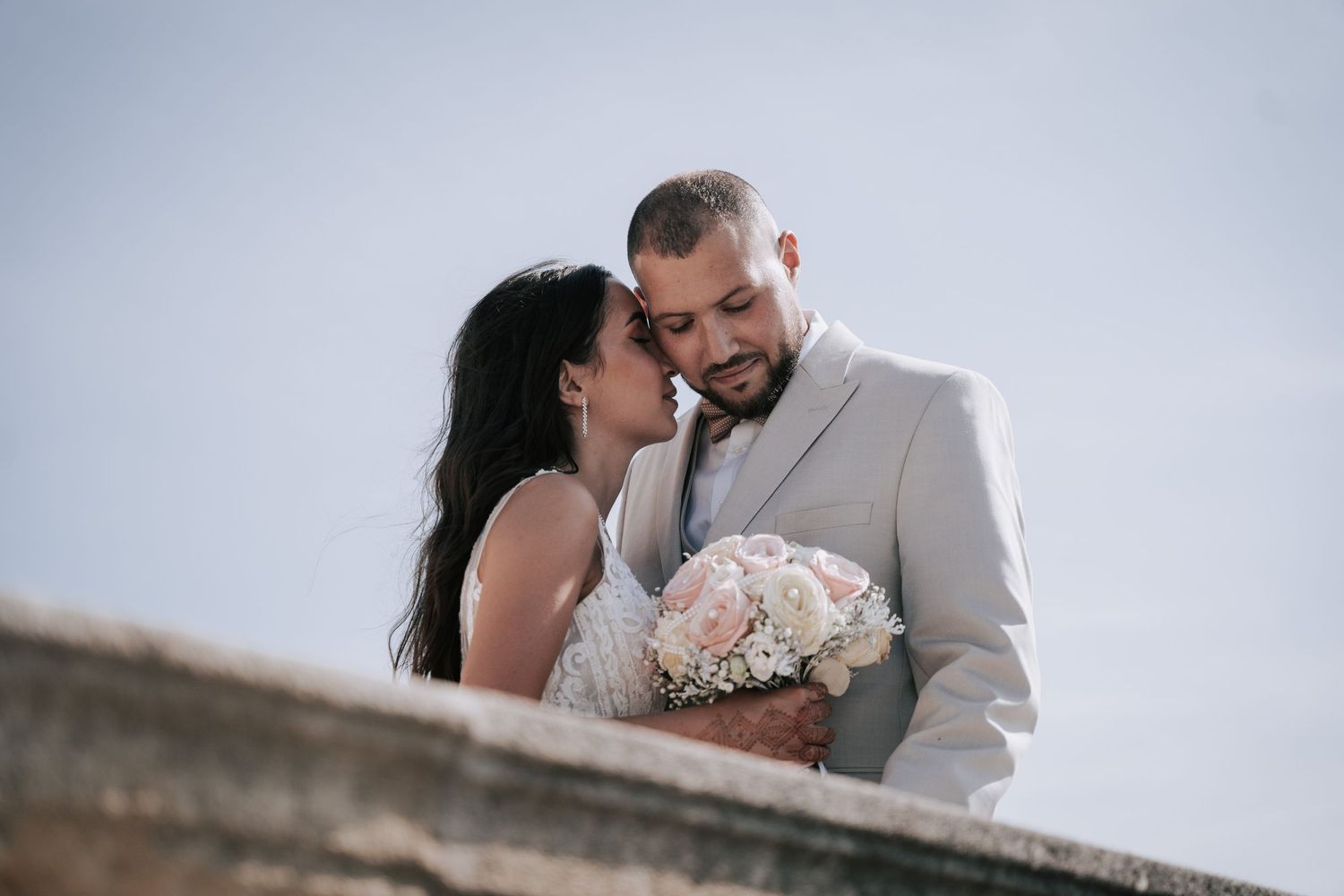 Bride and groom share a romantic moment holding a white bouquet against a blue sky.
