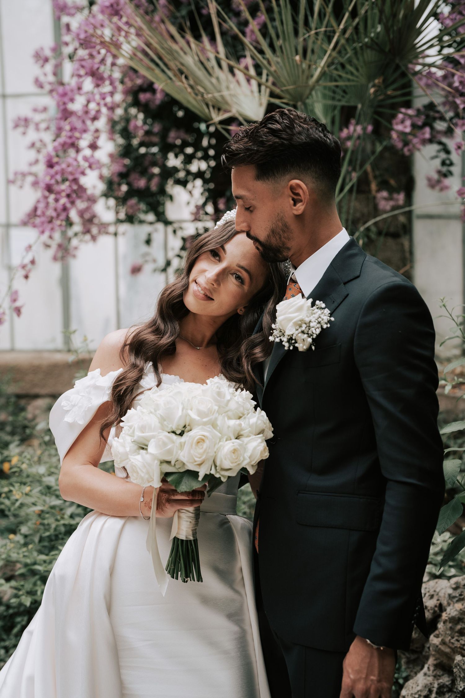 Groom in dark suit kisses bride on the head as she holds a white rose bouquet in a garden.