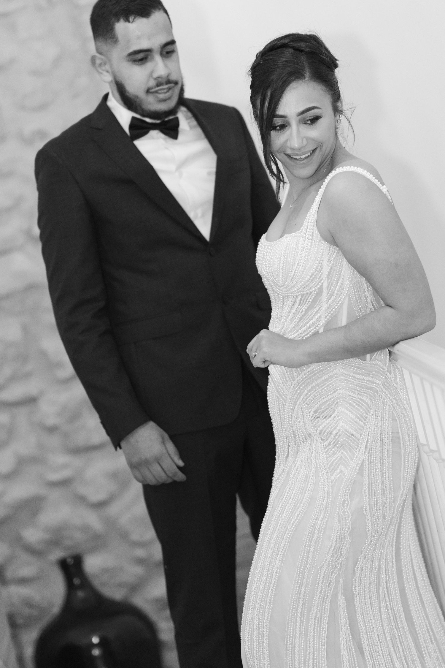 Black and white wedding portrait of groom standing behind smiling bride in elegant beaded gown near stone wall.