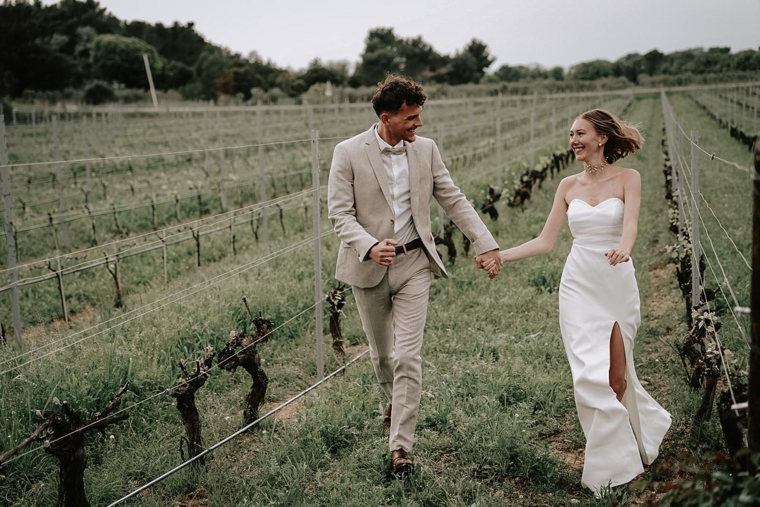 Happy bride and groom walking hand in hand through a lush vineyard on their wedding day.