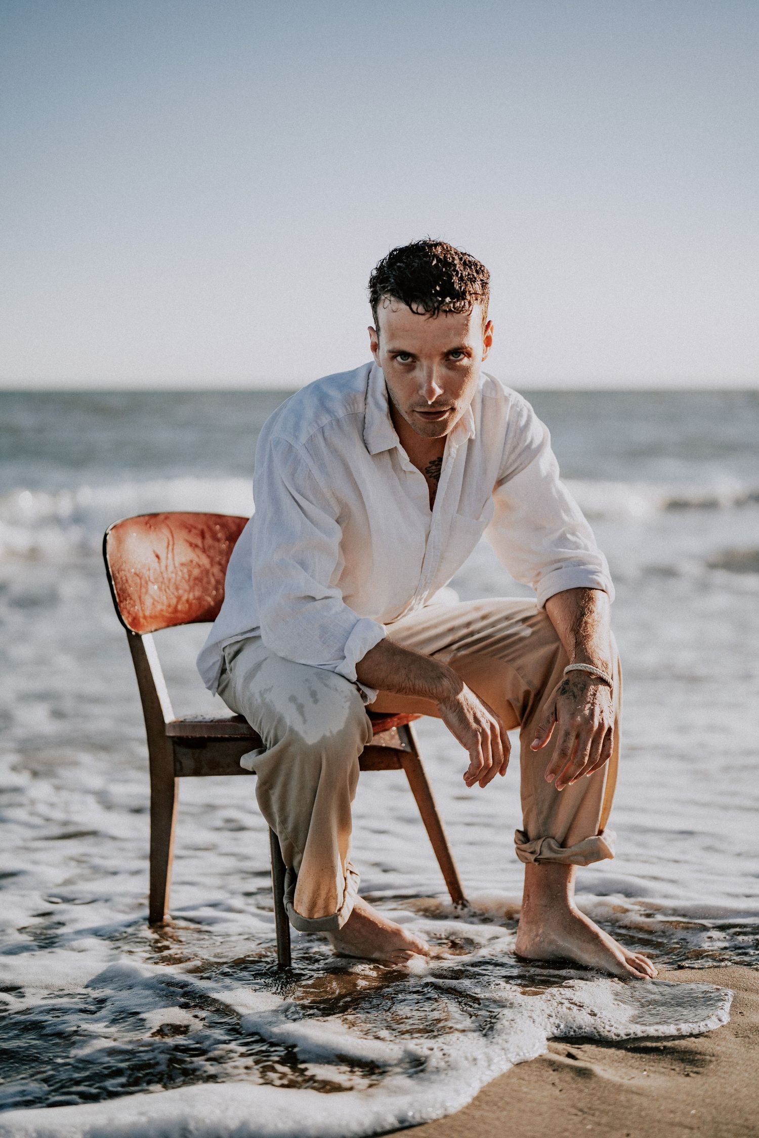Man in wet white shirt sitting on wooden chair in ocean waves on beach.