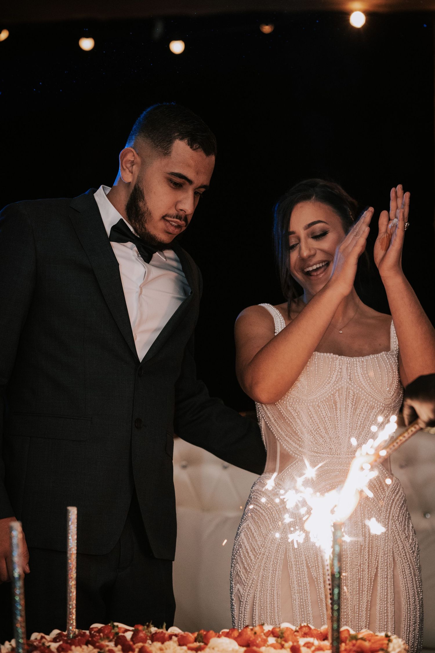 Bride and groom laughing together as wedding cake sparkler ignites during their evening reception celebration.