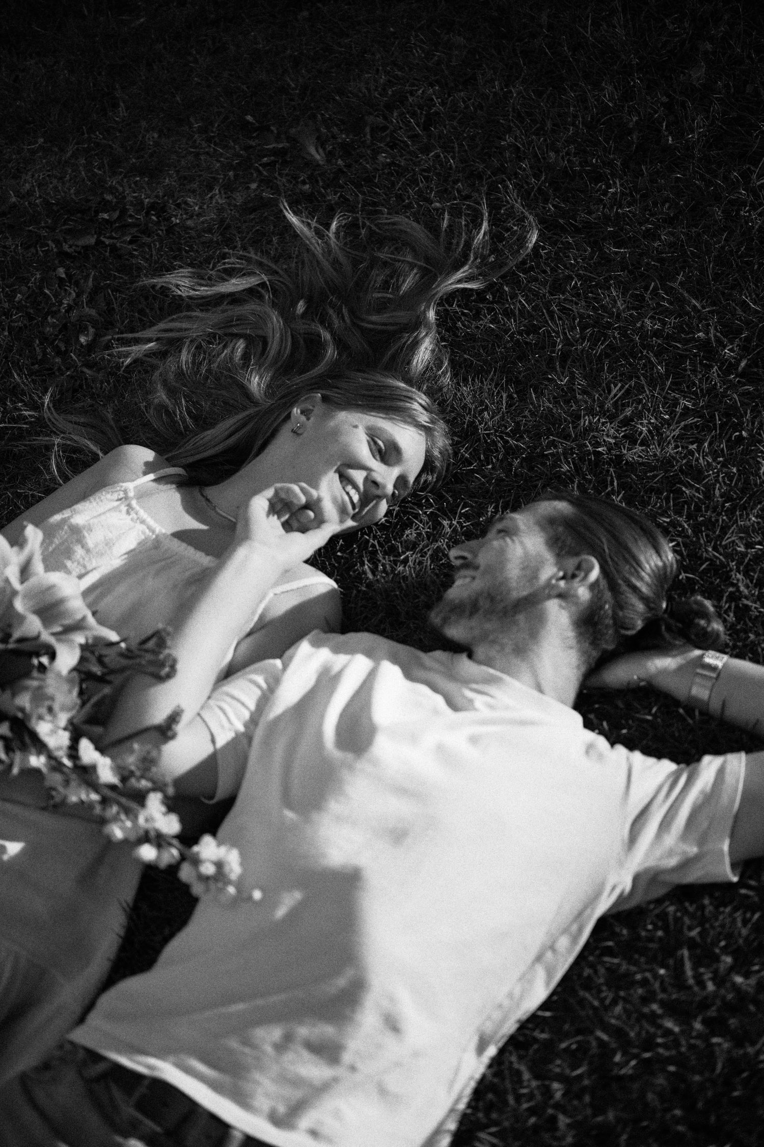 Black and white photo of a smiling couple lying on grass together in sunlight.