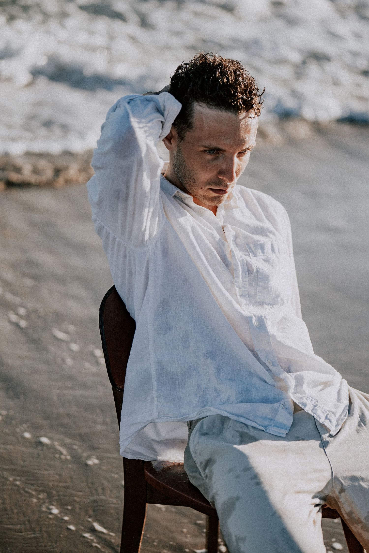 Man in wet white shirt posing on wooden chair on sandy beach near ocean.
