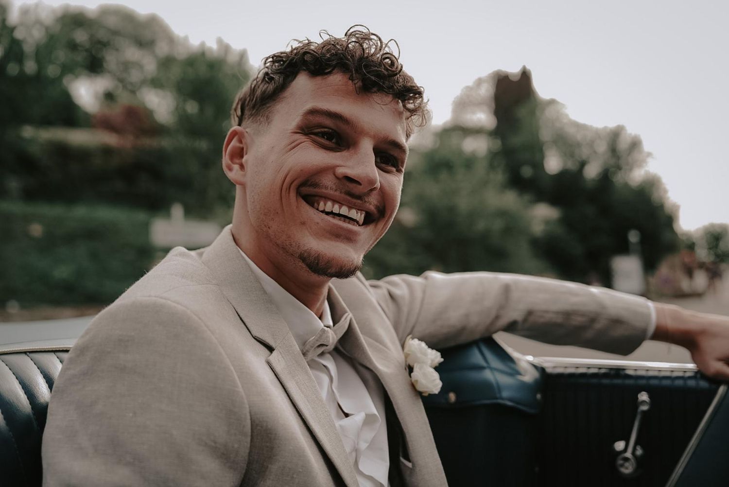 Handsome groom in beige suit smiling in a vintage convertible car on his wedding day.
