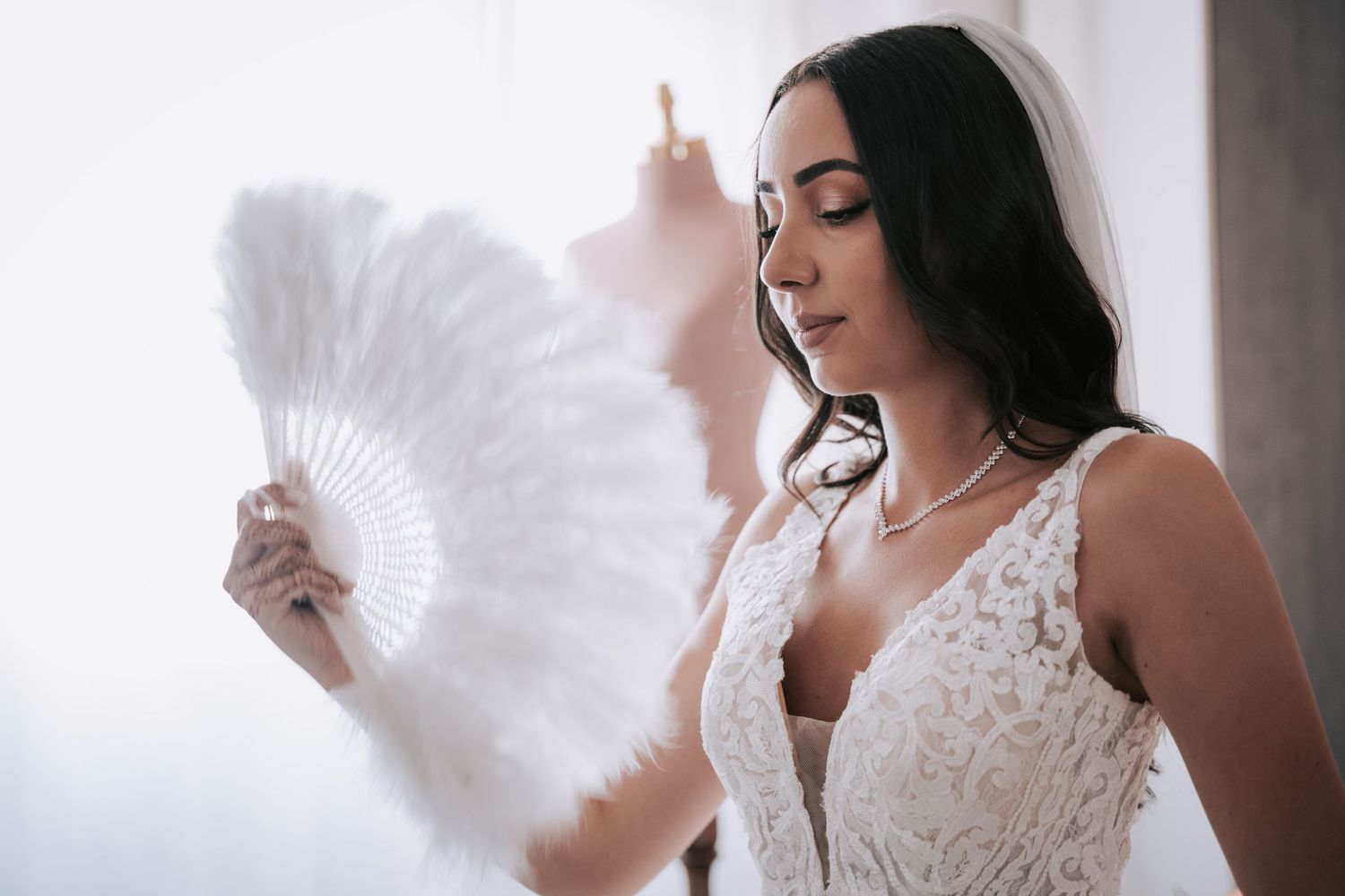 Elegant bride in lace wedding gown holding a white feather fan near a window with soft natural light.
