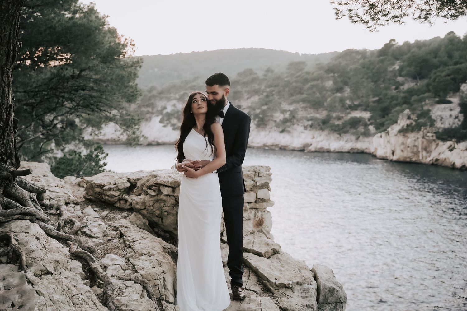 Bride and groom embrace on rocky cliffs overlooking a serene coastal bay with lush green hills.