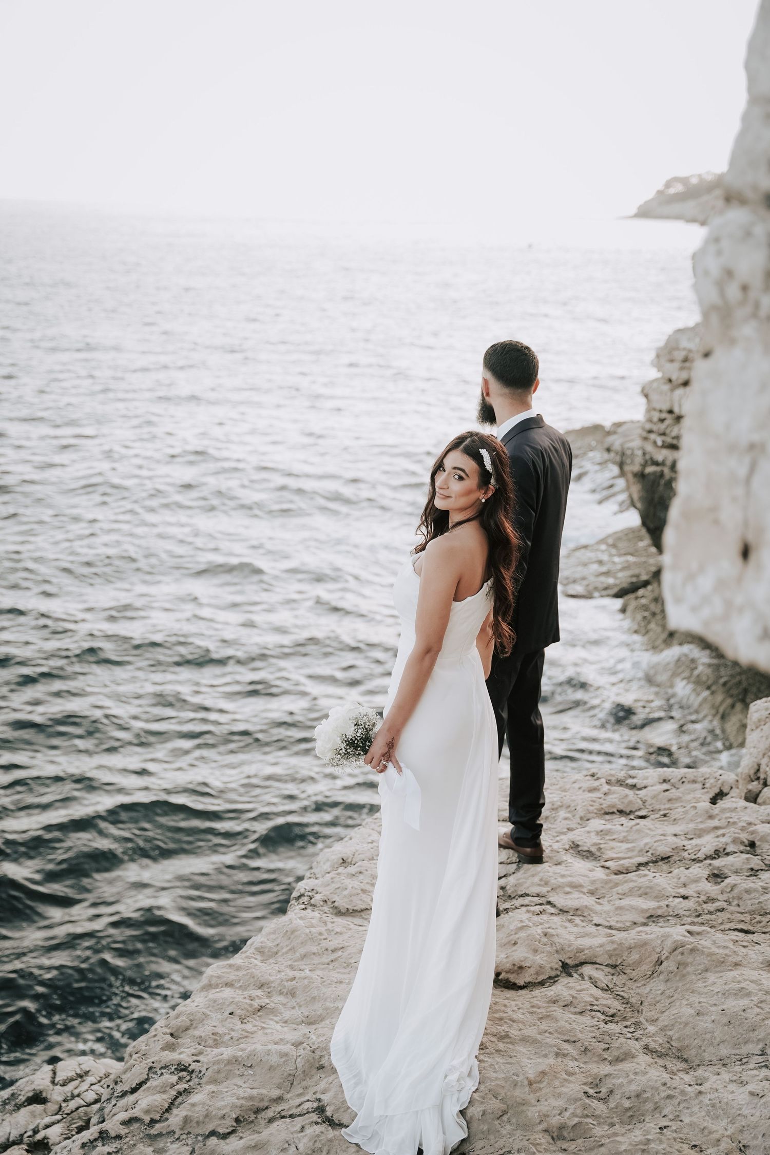 Bride in flowing white gown stands on rocky coastal cliffs with groom overlooking the sea.