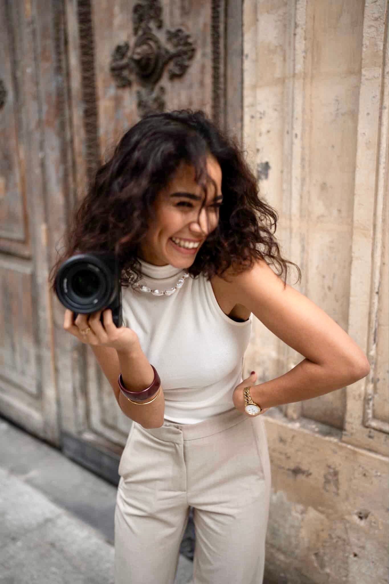 Smiling woman with curly hair holding a camera, wearing white outfit and jewelry, posing outdoors near ornate wooden door.