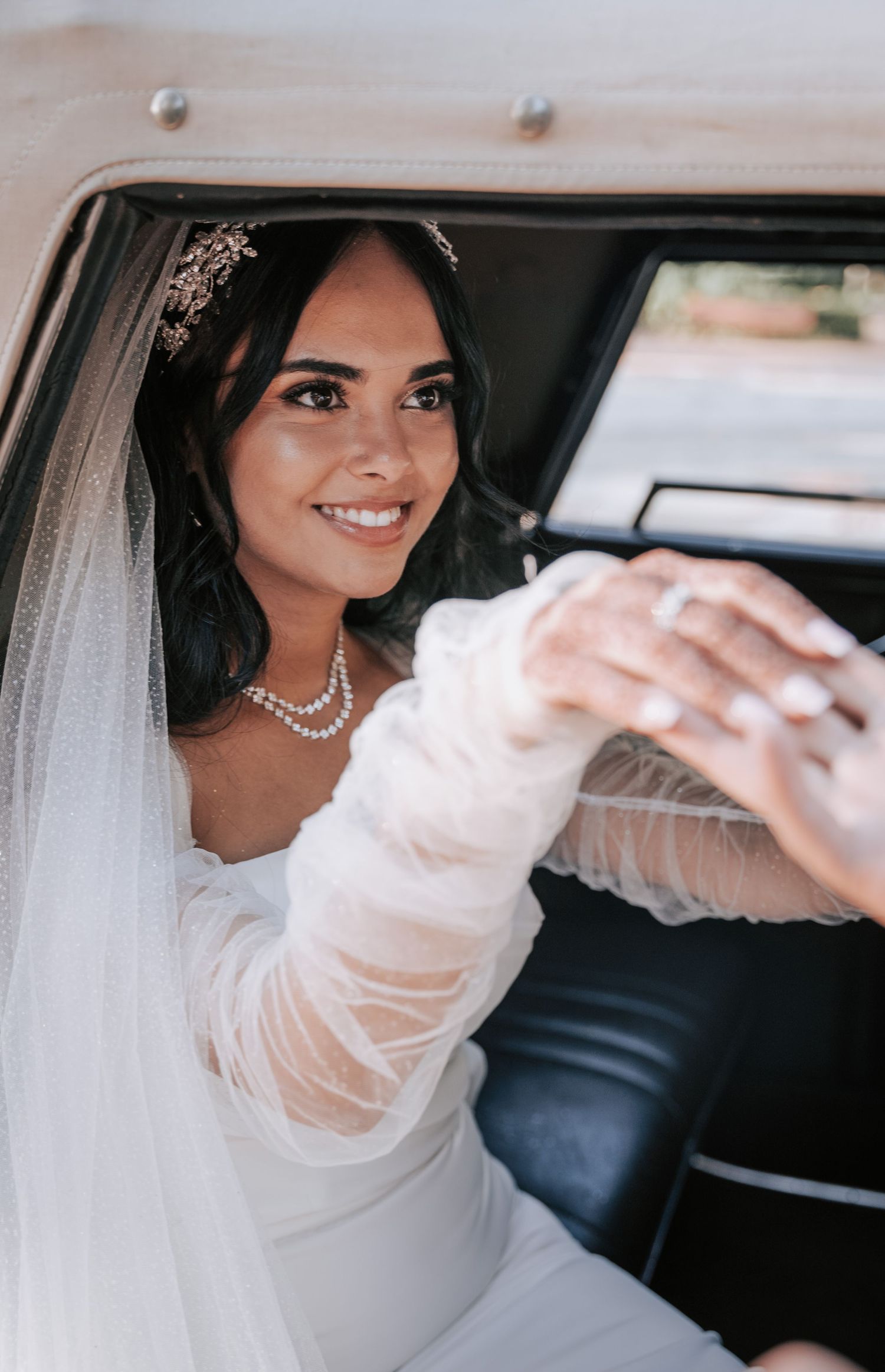 Smiling bride in white gown and veil sits in vintage car, wearing pearl necklace and floral hair accessory.