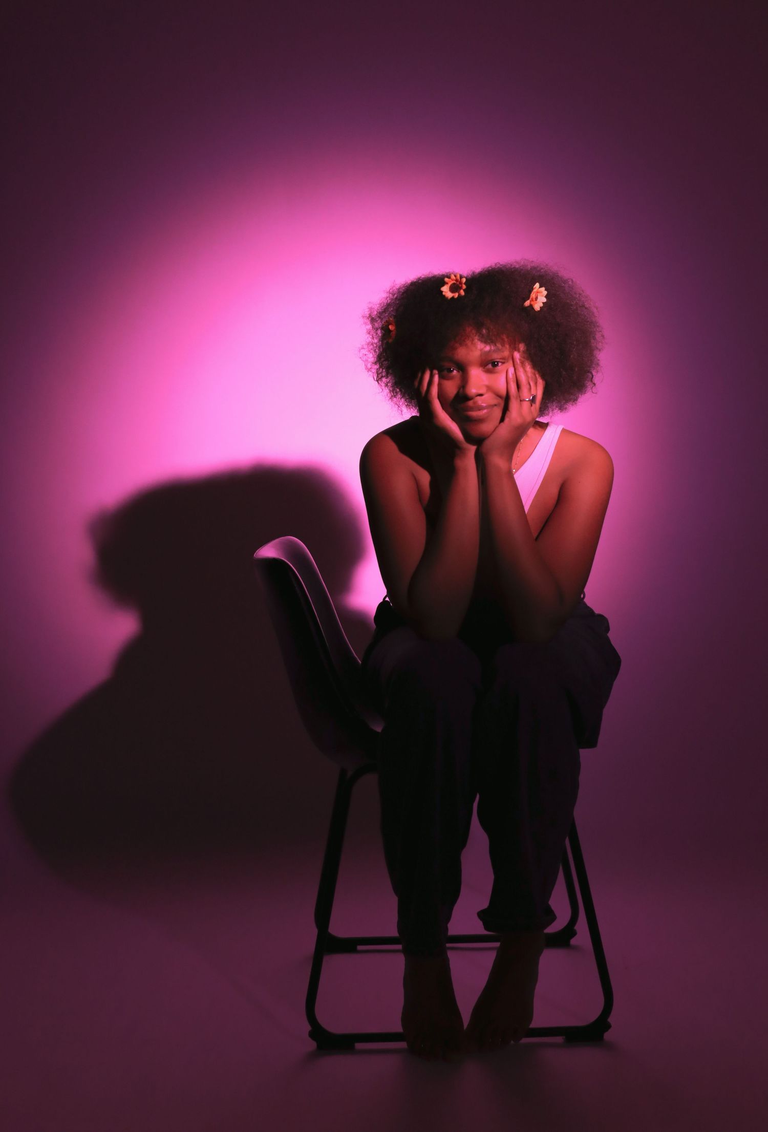 Young woman with afro puffs sits on chair, hands on cheeks, smiling in pink-purple studio lighting.