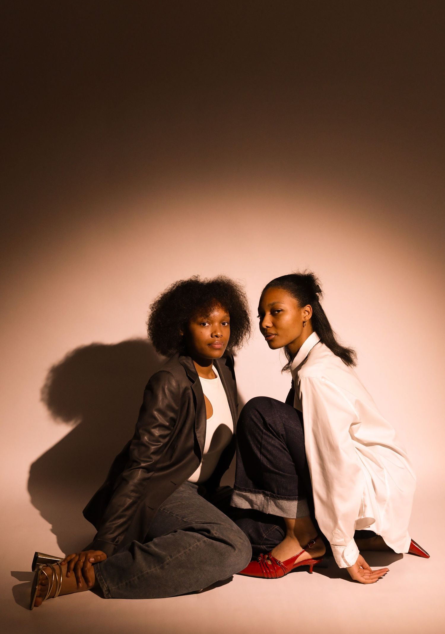 Two Black women models pose seated on floor, one in dark blazer with afro, other in white shirt with sleek ponytail.