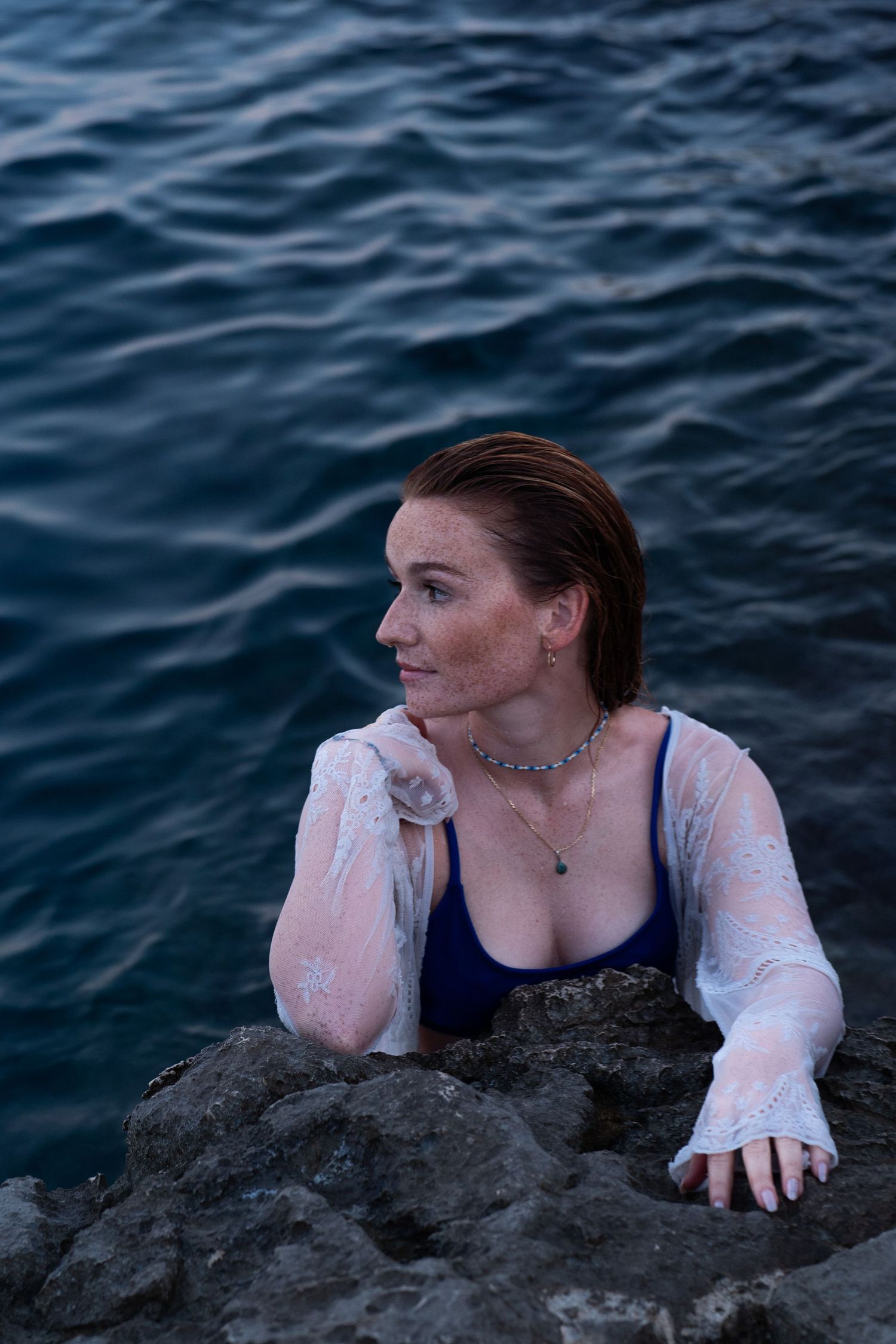 Woman in blue bikini and white lace top resting on rocky shoreline with ocean backdrop.