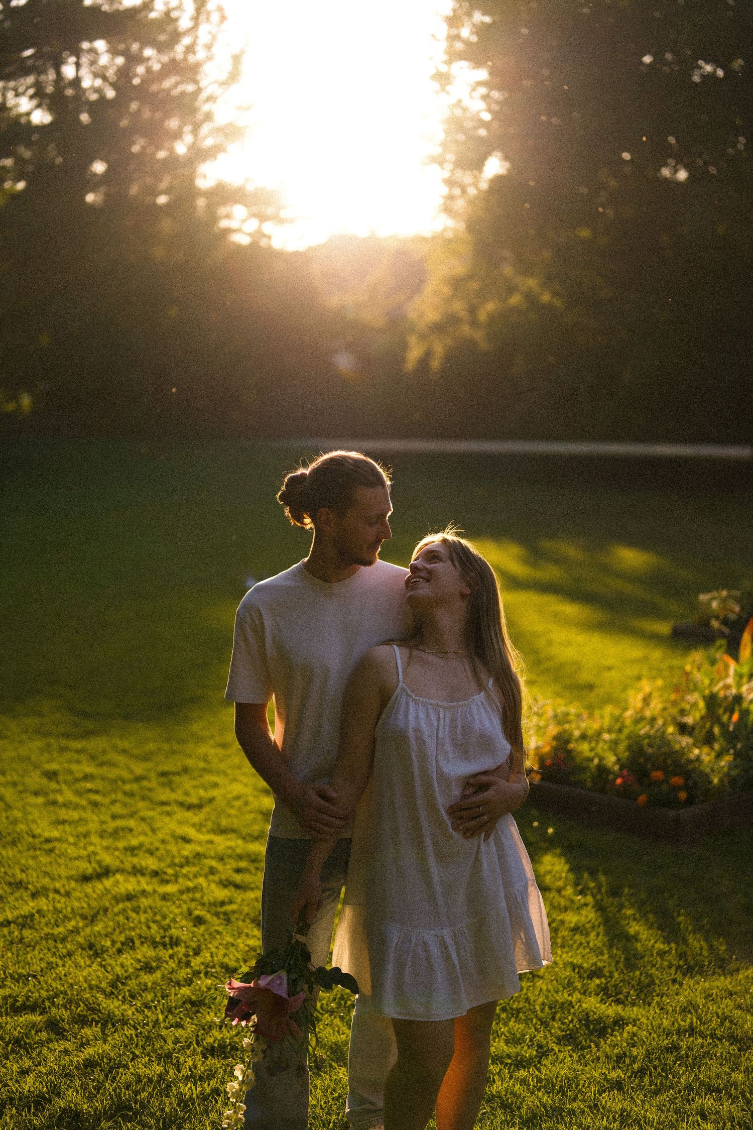 Romantic couple laughing and embracing on a green lawn during a beautiful golden hour sunset in a park.