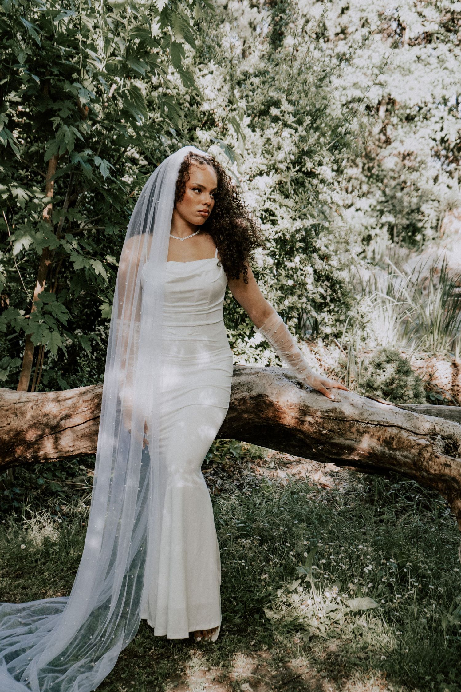 Bride in fitted white mermaid gown with pearl veil leans gracefully against a fallen log in a garden.