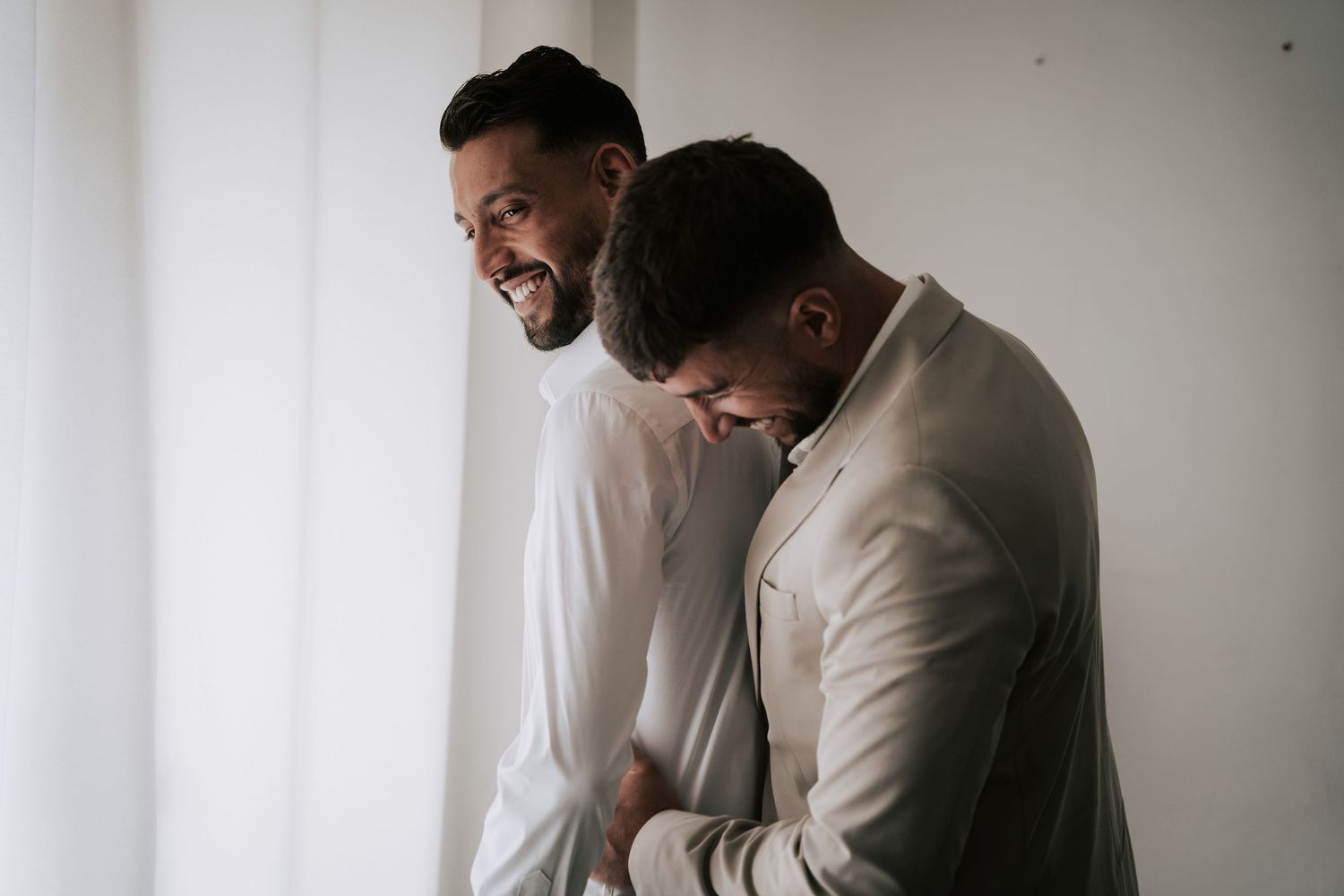 Two men in suits laughing together during wedding preparations in a bright white room.
