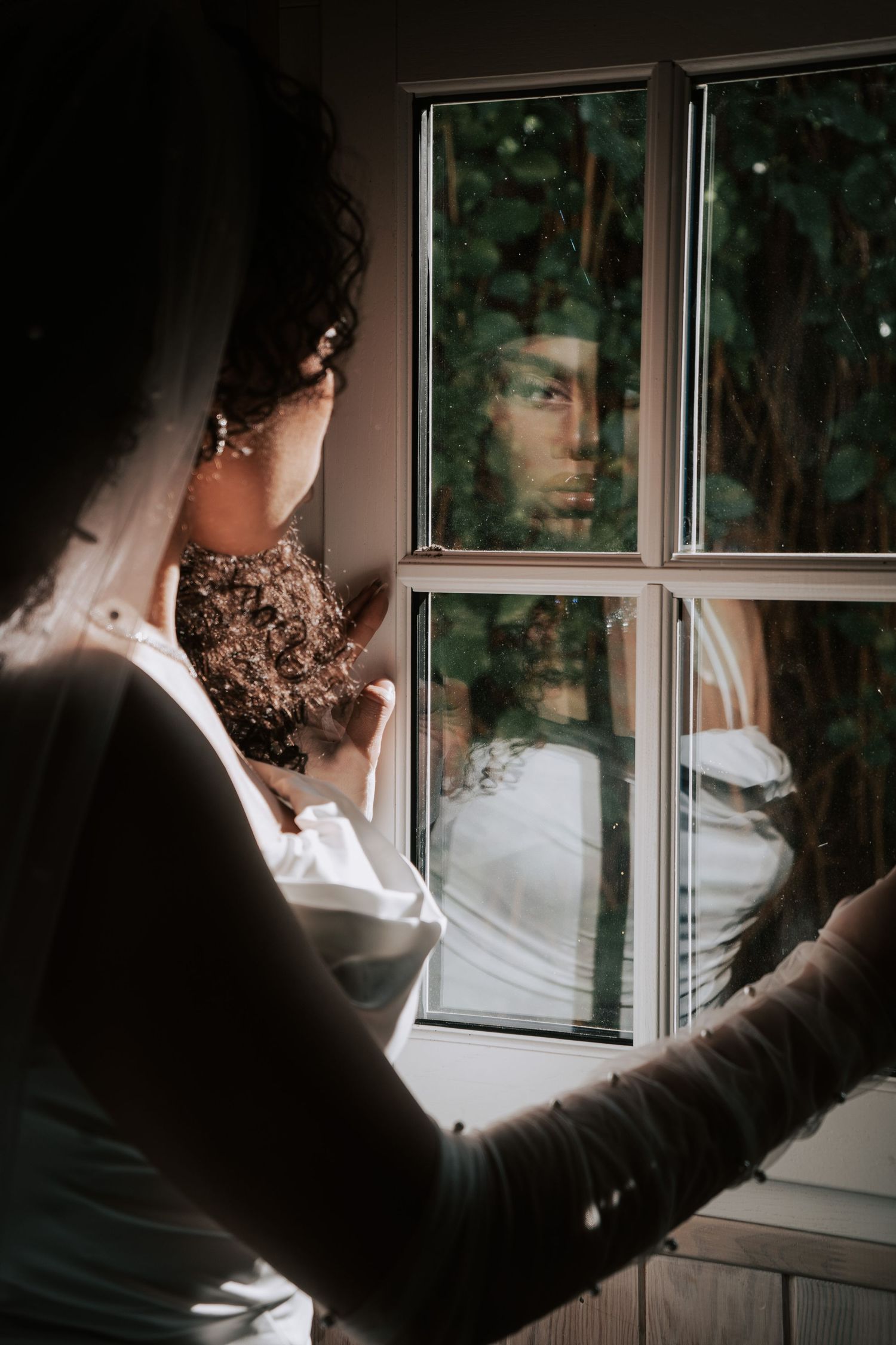 Bride in white gown and veil gazes at her reflection in a multi-pane window surrounded by lush greenery.