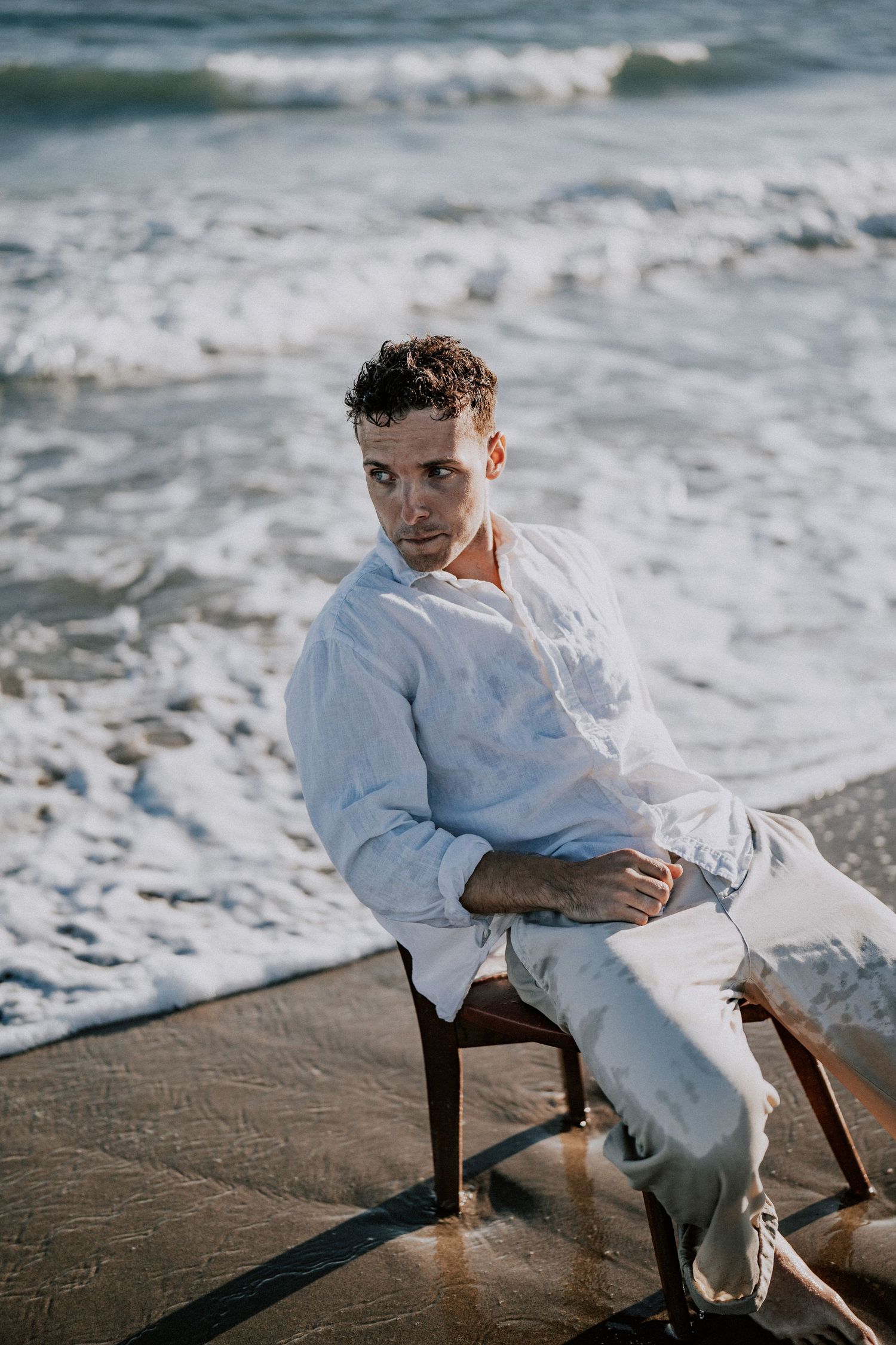 A handsome young man in a white shirt and light pants sits on a chair near crashing ocean waves.