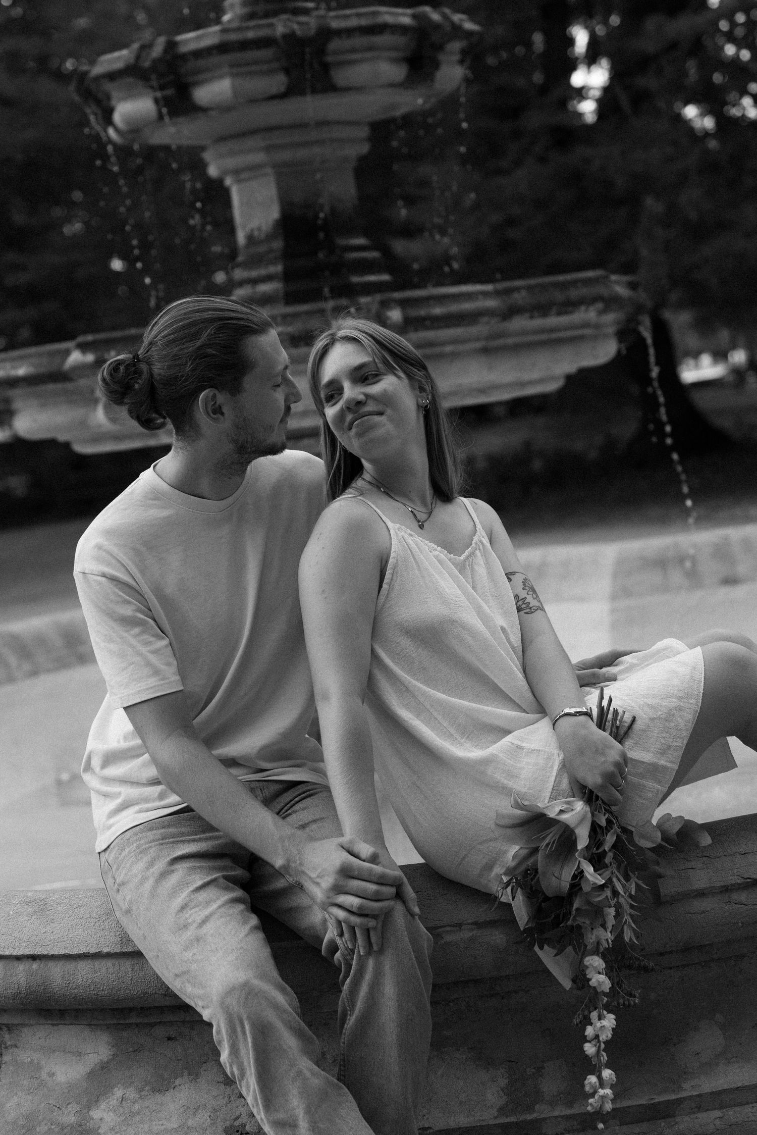 Black and white photo of a couple sitting on a fountain edge, woman holding flowers, man leaning in to kiss her cheek.